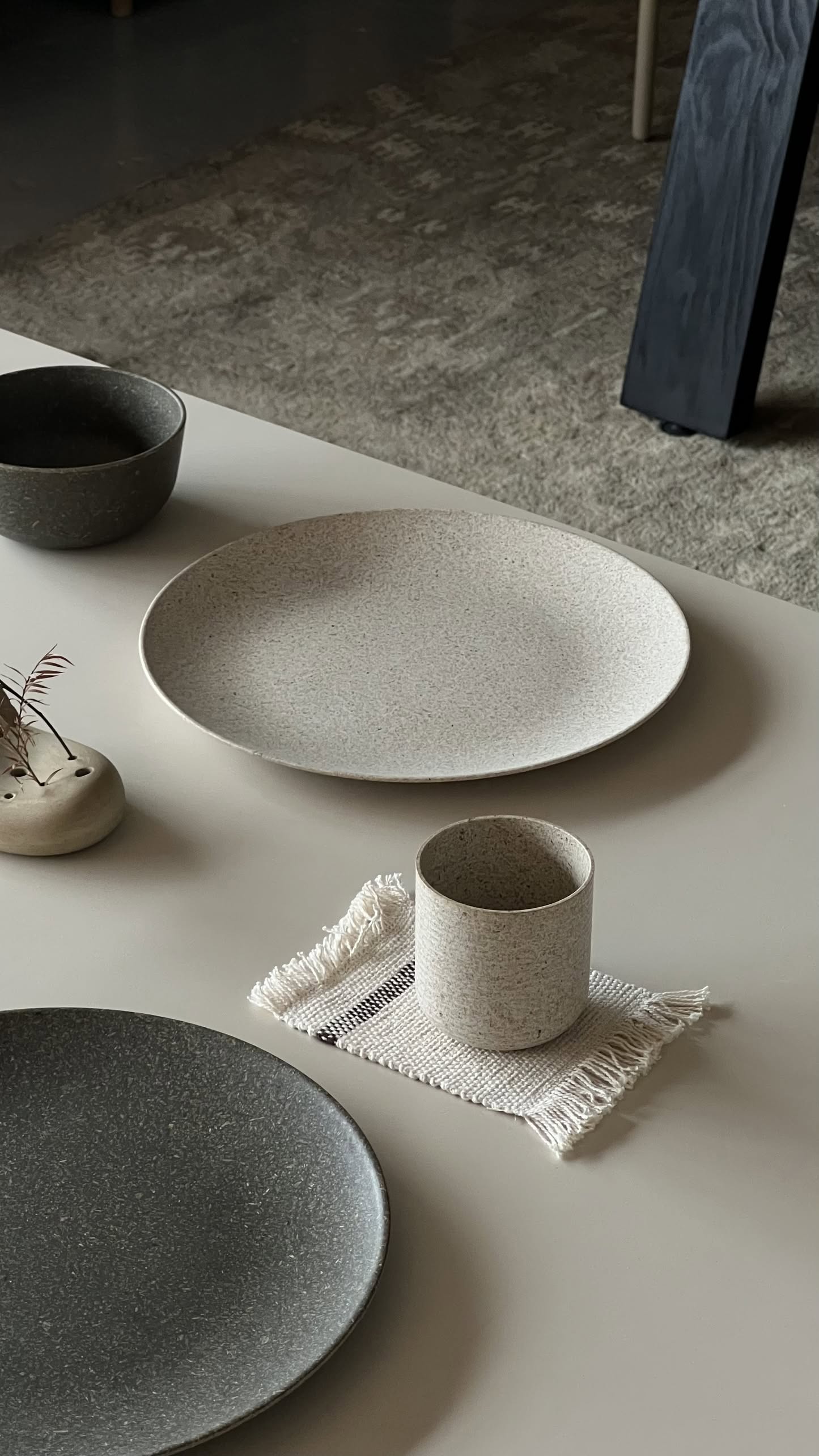 Minimalist table setting with the eco-conscious Pass It On Dining Plate L, Rice Husk by Pass It On, paired with neutral-toned ceramics, a dark bowl, a small vase of dried foliage, and a cup on a fringed cloth for sustainable dining.