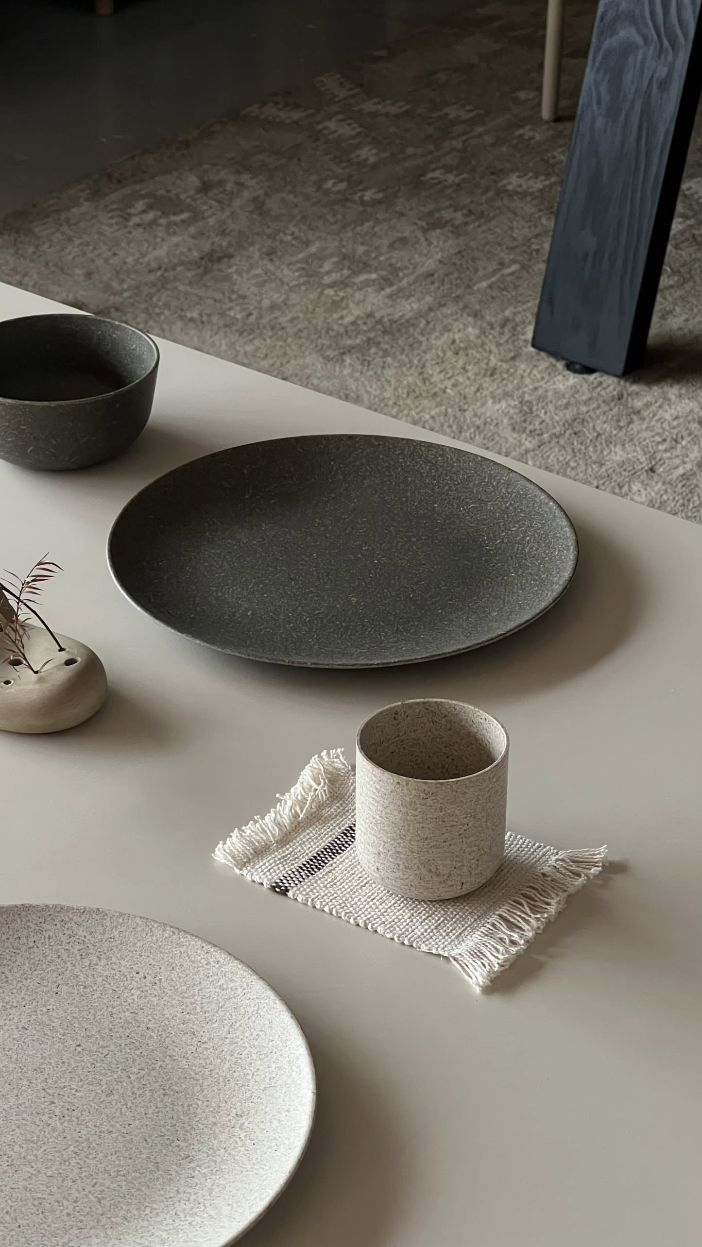 A minimalist tabletop features the Pass It On Dining Plate L made from rice husk, neutral-toned, paired with a cup on a fringed cloth, eco-conscious tableware, and a small vase with dried foliage on a light-colored surface.