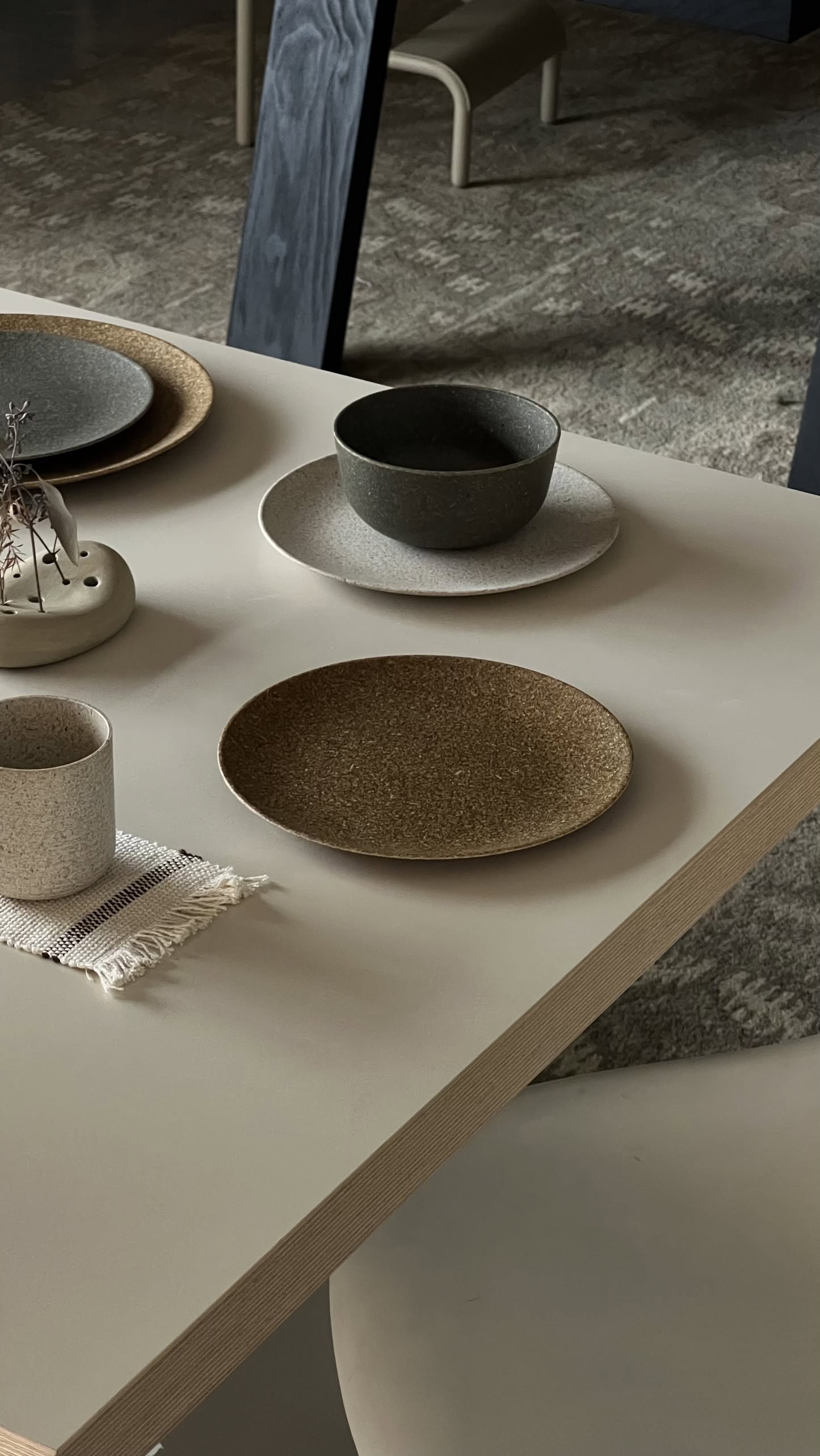 A minimalist dining table set featuring the Pass It On Dining Plate M (Rice Husk) by Pass It On, paired with a neutral-toned bowl, cup, small woven mat, and a simple vase with dried twigs—earthy muted tones for modern design lovers.
