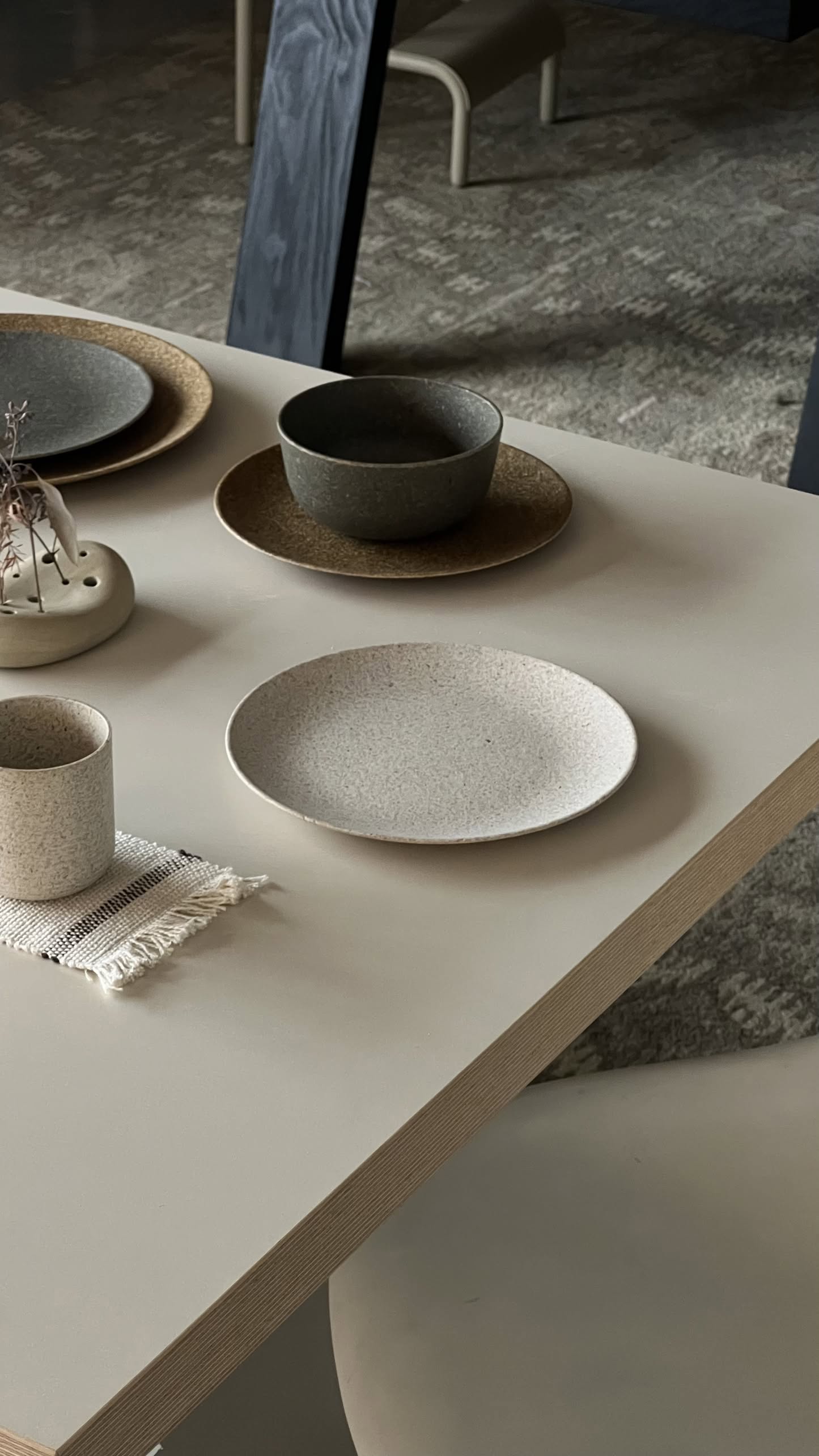 A minimalist table setting with the Pass It On Dining Plate M (Rice Husk) by Pass It On, paired with bowls, a cup on a woven mat, and a ceramic vase of dried twigs—an eco-conscious and modern design statement on a light-colored table.