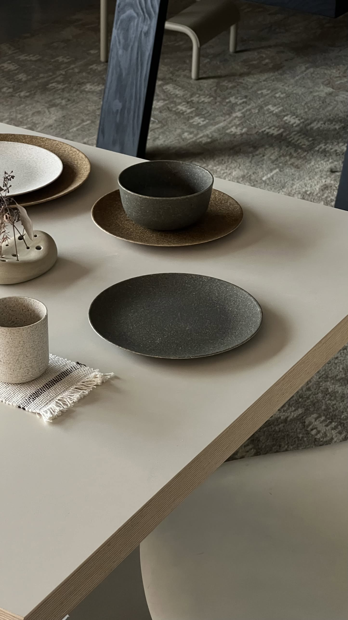 A minimalist table setting with neutral-toned plates, bowls, and a cup features the Pass It On Dining Plate M, Rice Husk by Pass It On on a light table. A small vase with dried flowers and a fabric coaster add modern design, with a softly blurred background.