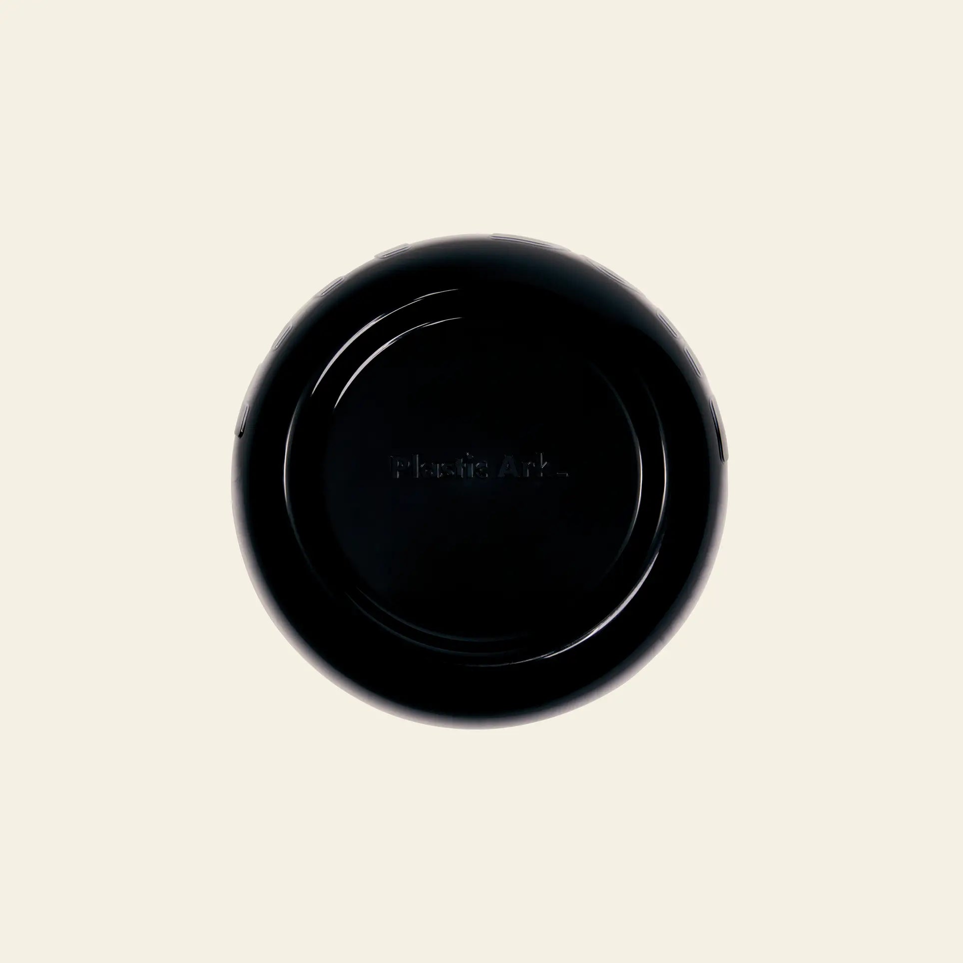 A black, round Plastic Ark Fat Bowl from Plastic Ark is seen from above on an off-white background. Plastic Ark is faintly visible at the center, highlighting its recycled plastic construction and customizable design.