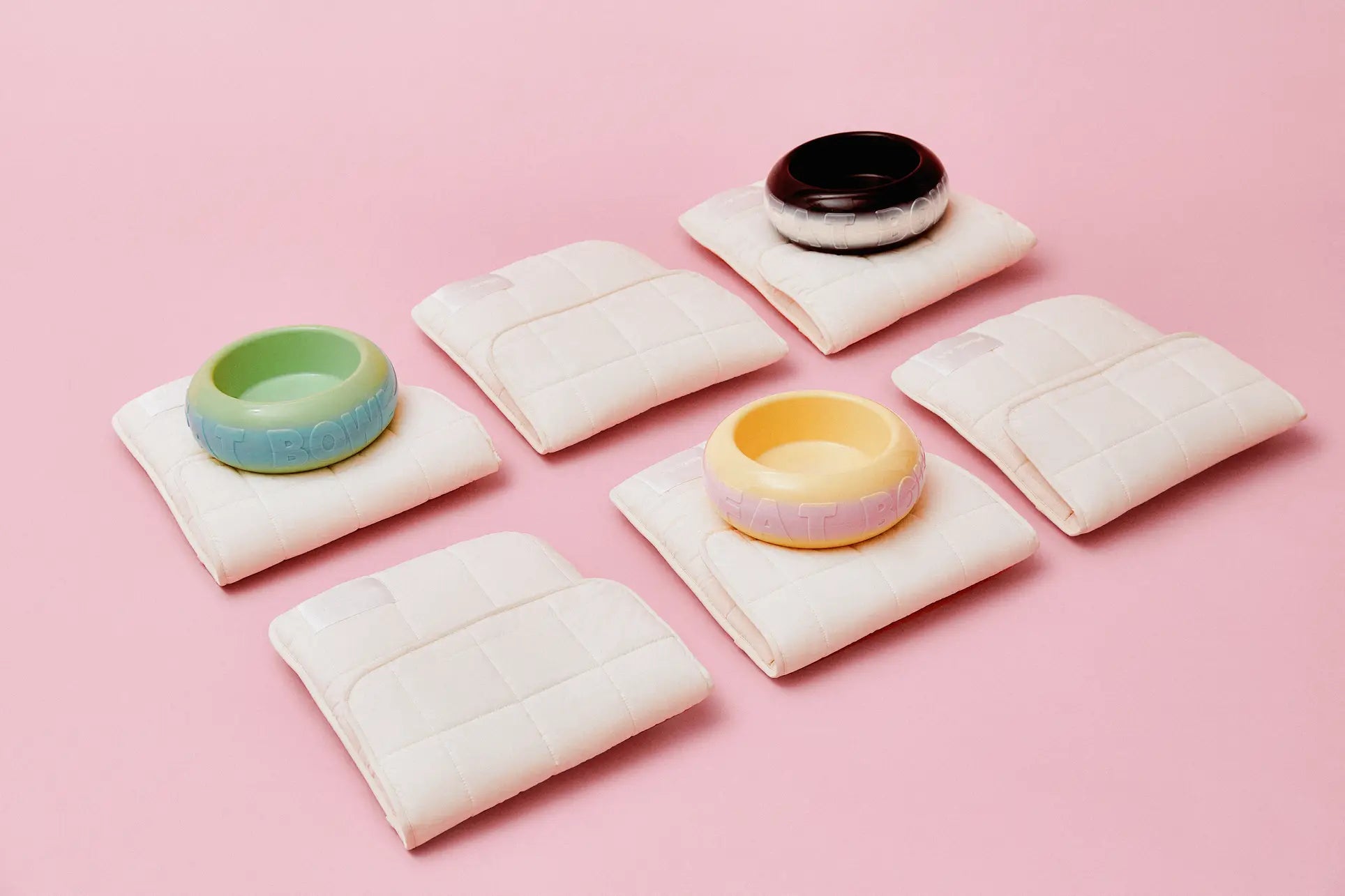 Six folded cream-colored quilted pads are on a pink background. The top three hold Plastic Ark Fat Bowls (green, black, yellow) by Plastic Ark, made from recycled plastic. The bottom three pads are empty.