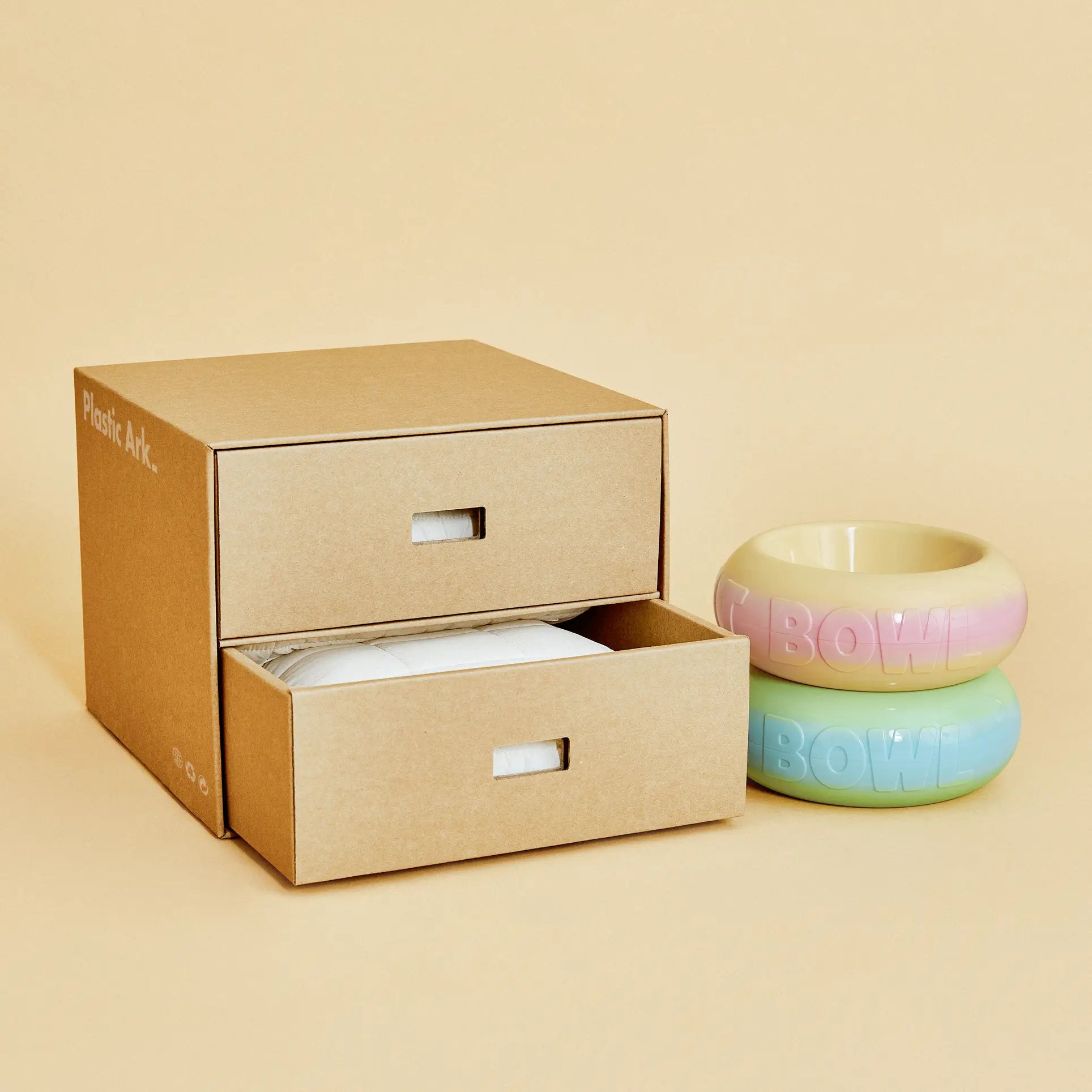 A brown cardboard box with two drawers, one half-open with folded white fabric inside, sits beside two stacked Plastic Ark Fat Bowls by Plastic Ark in pastel colors against a beige background.