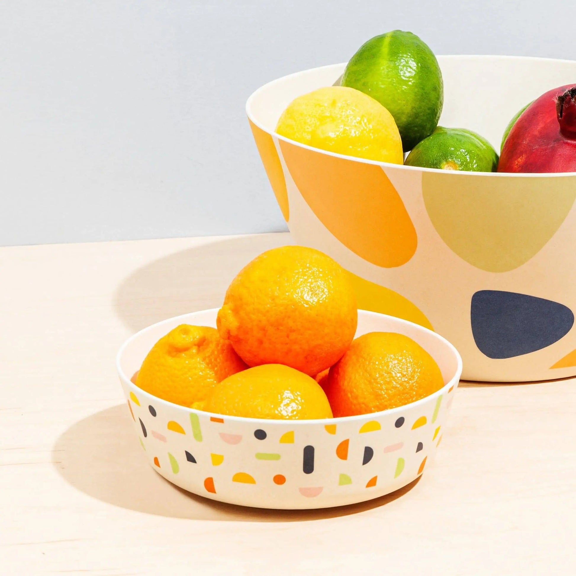 Two Poketo Bamboo Bowls by Poketo sit on a light surface; the front bowl is filled with oranges, and the larger back bowl holds a lemon, limes, and a red apple. These eco-friendly bowls feature vibrant, abstract patterns inspired by outdoor adventure.