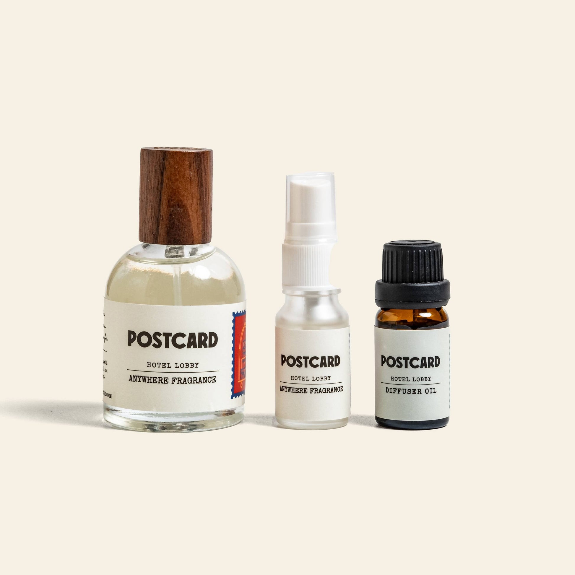 Three Postcard bottles are lined up: a wooden-capped spray, white plastic spray, and small amber diffuser oil. Each features Hotel Lobby and Anywhere Fragrance text, evoking luxury with this bestselling Postcard scent.
