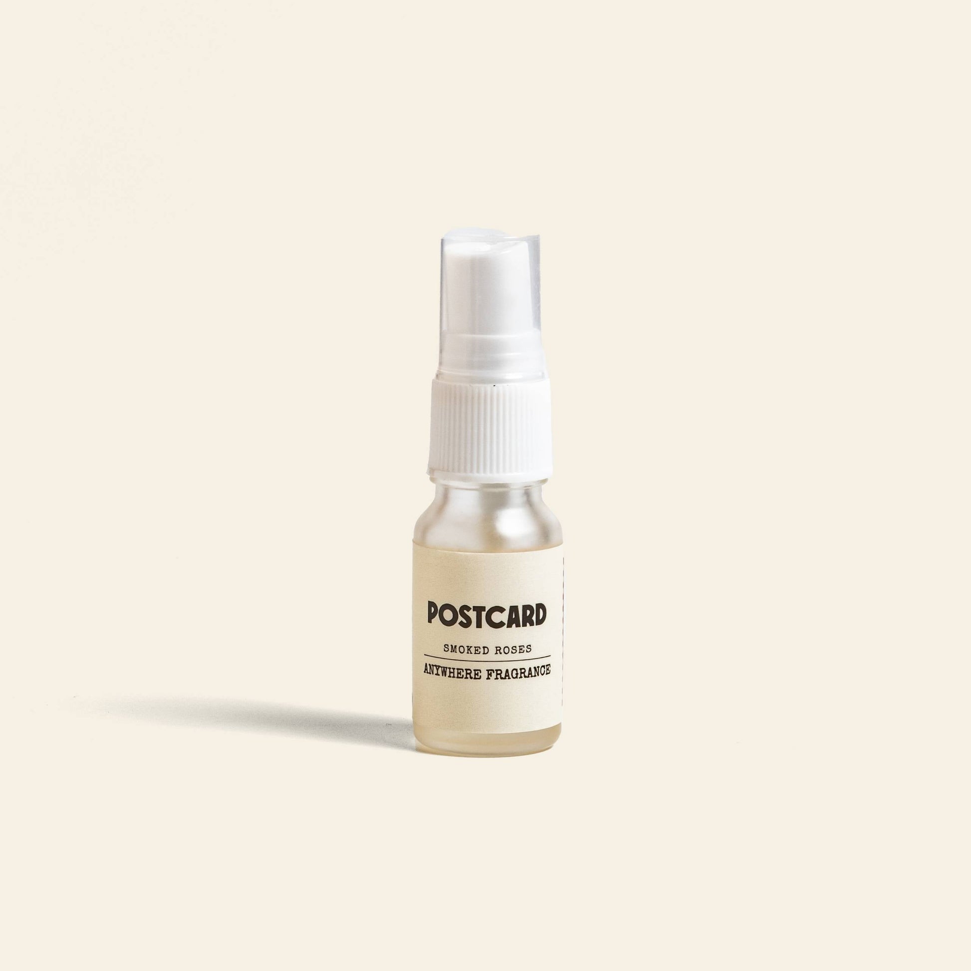 A 10ml Postcard Anywhere Fragrance in Smoked Roses by Postcard, with a white cap, rests on a beige background, evoking subtle amber and rose notes.