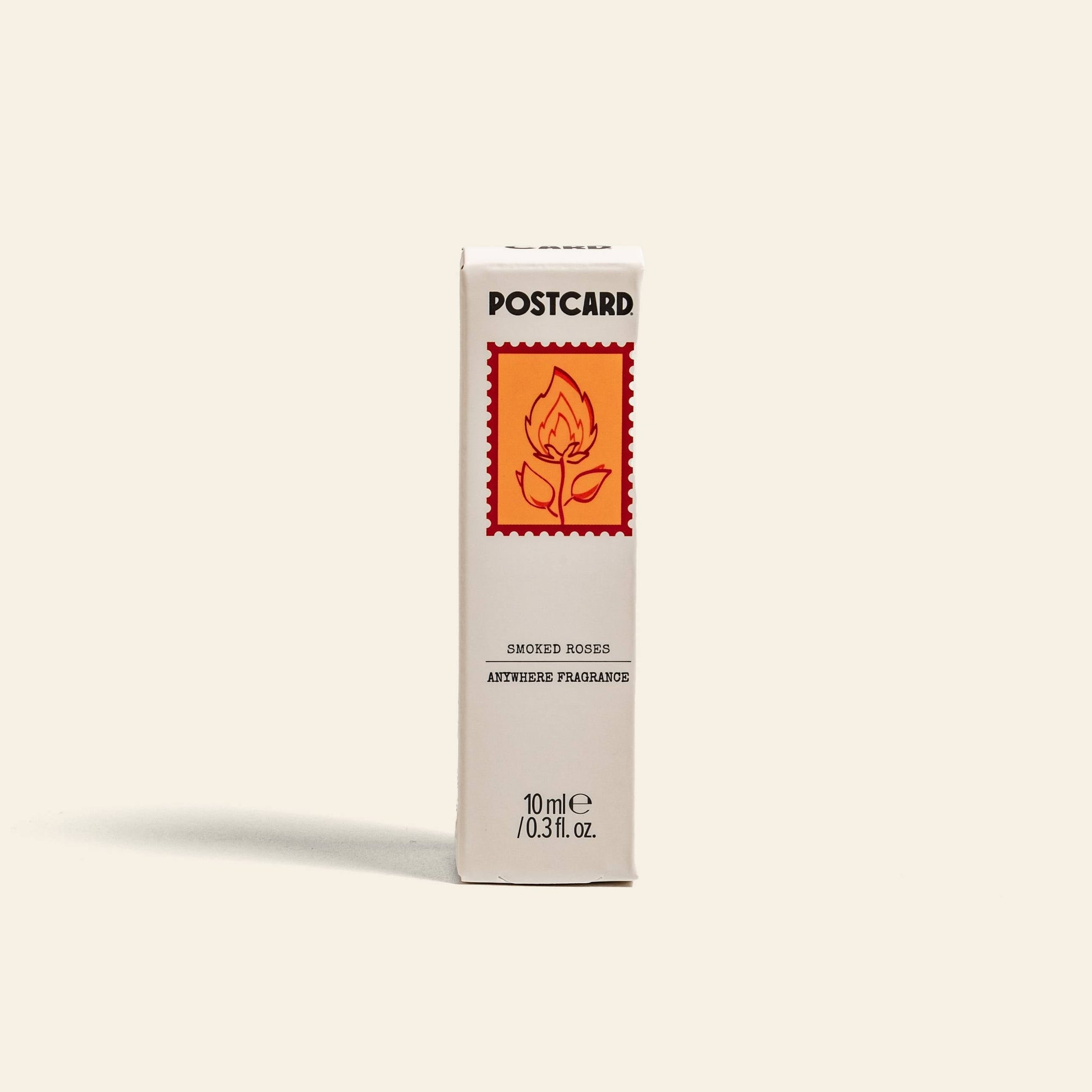 A white rectangular box labeled Postcard features an orange rose stamp and highlights the Smoked Roses scent—an amber and rose blend, in a 10ml (0.3 fl. oz.) anhydrous perfume spray—from Postcard Anywhere Fragrance.