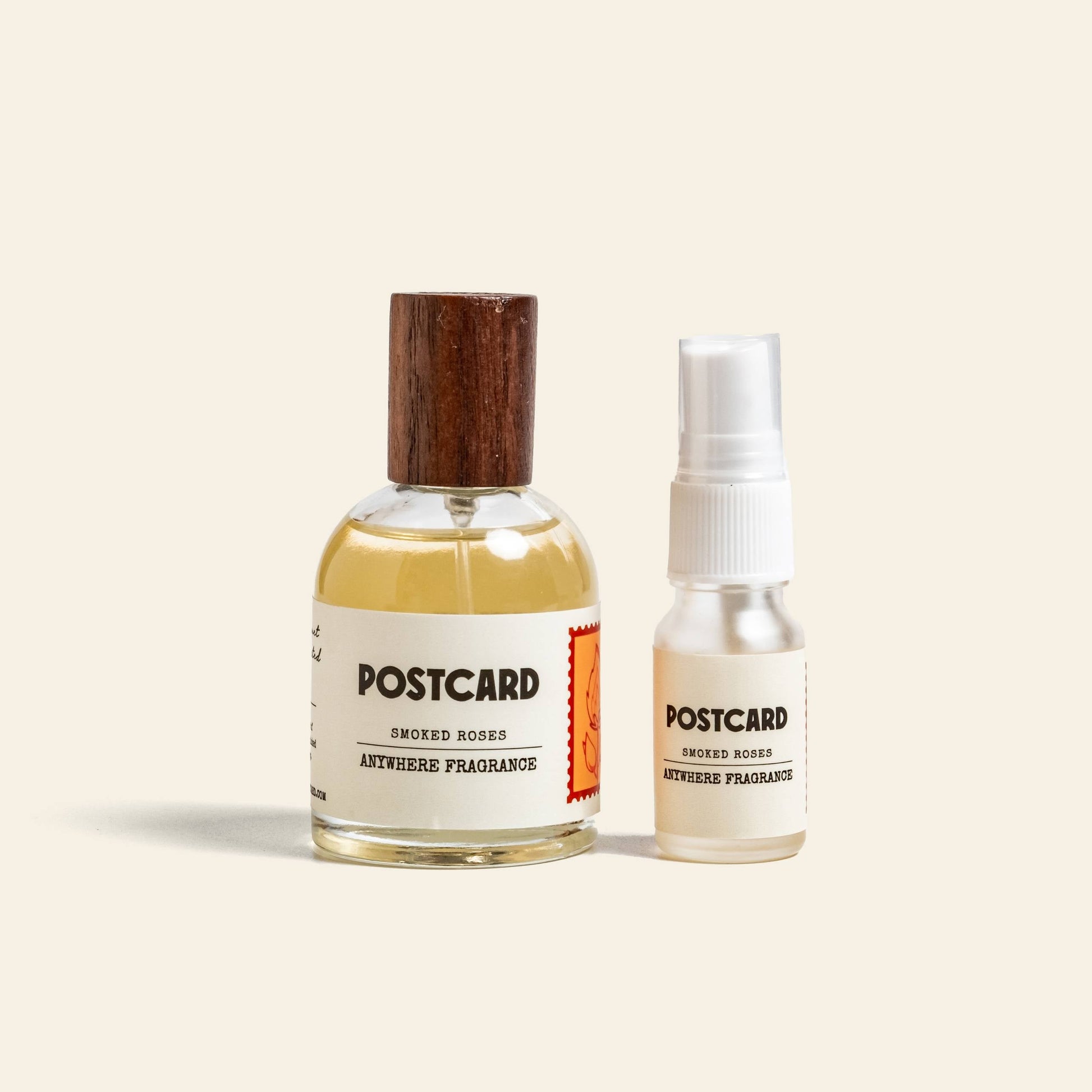 Two bottles of Postcard Anywhere Fragrance, 10ml - Smoked Roses by Postcard, with warm amber and rose notes, sit side by side—a larger bottle with a wooden cap and a smaller spray—on a light beige background.