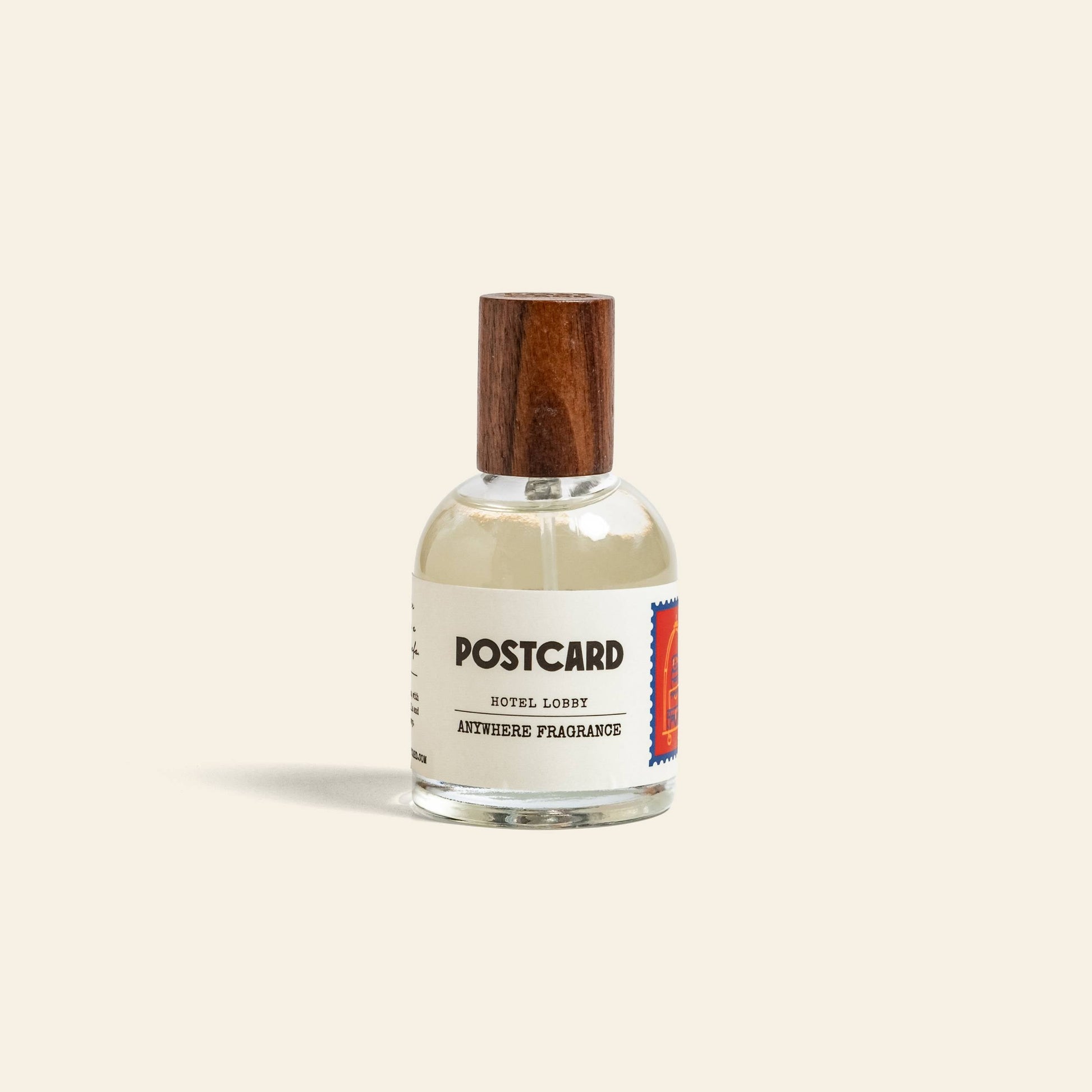A clear glass bottle of Postcard Anywhere Fragrance, 50ml - Hotel Lobby, with a wooden cap and a white label with blue and red stamp design, stands on an off-white background, evoking hotel luxury in this bestselling scent.