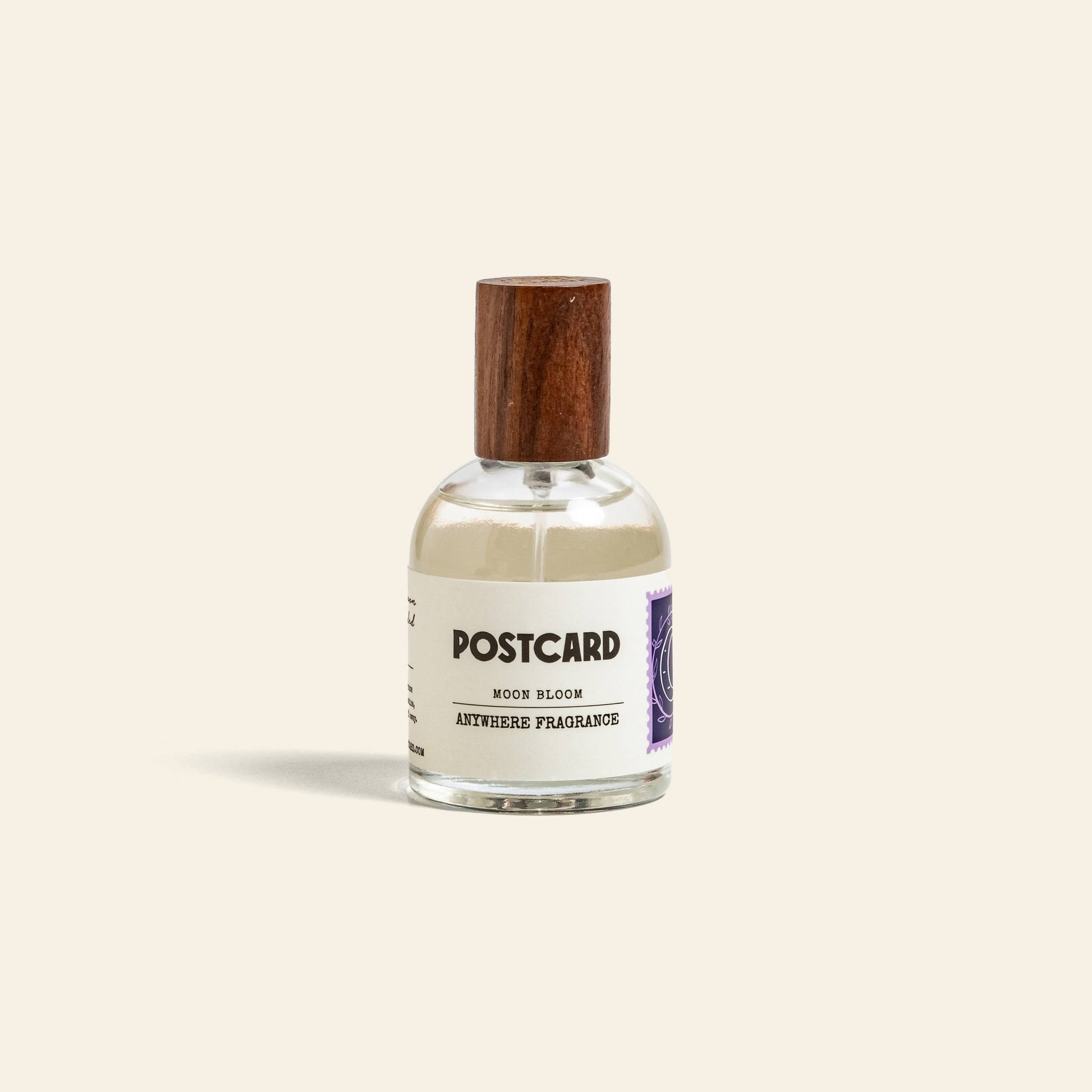 A clear 50ml Postcard Anywhere Fragrance—Moon Bloom—with a wooden cap, white label, and purple stamp graphic sits against a beige background, capturing dreamy scent notes in elegant glass.