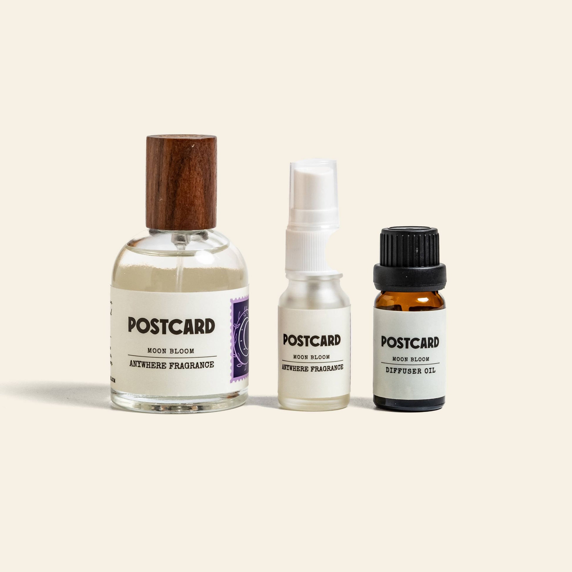 Three Postcard products are shown: a glass bottle with wooden cap, a small 50ml Postcard Anywhere Fragrance in Moon Bloom, and a dark glass dropper—each holding unique Scent Notes or diffuser oils.