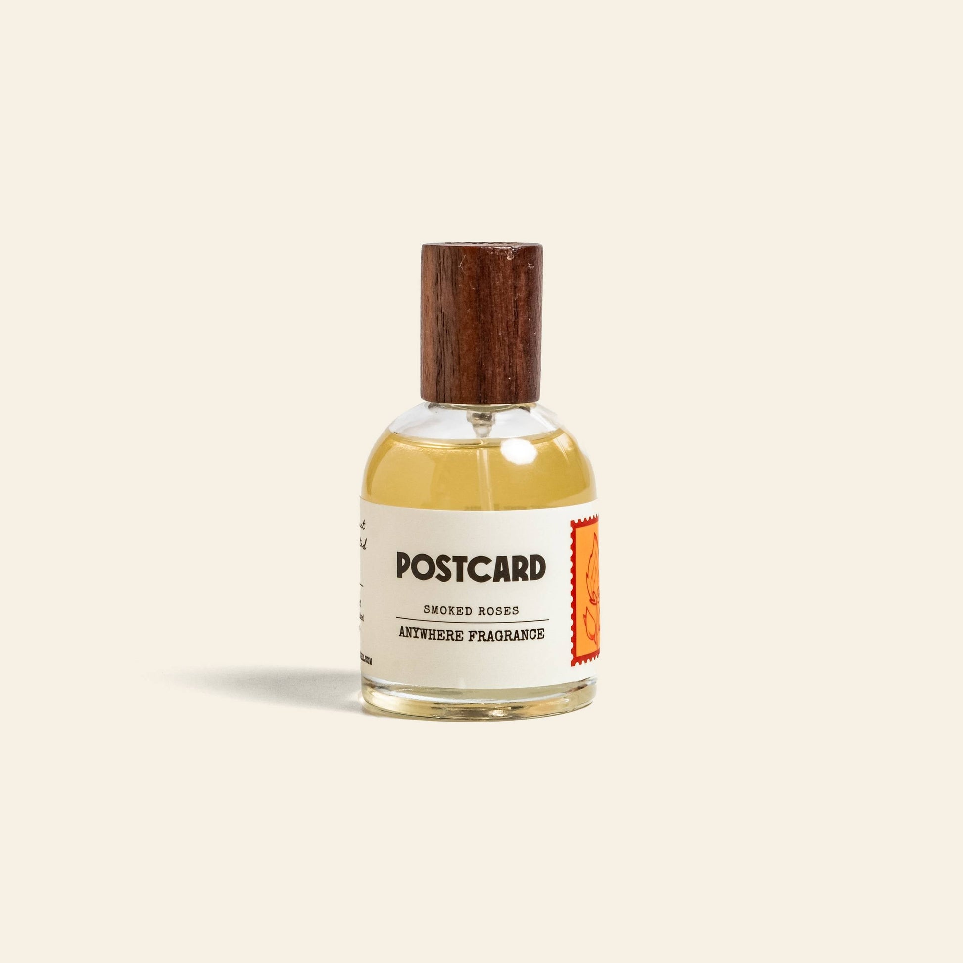 A clear glass bottle with a wooden cap holds Postcard Anywhere Fragrance, 50ml - Smoked Roses by Postcard. The label features a red stamp and hints at rose scent notes, with light amber liquid inside.