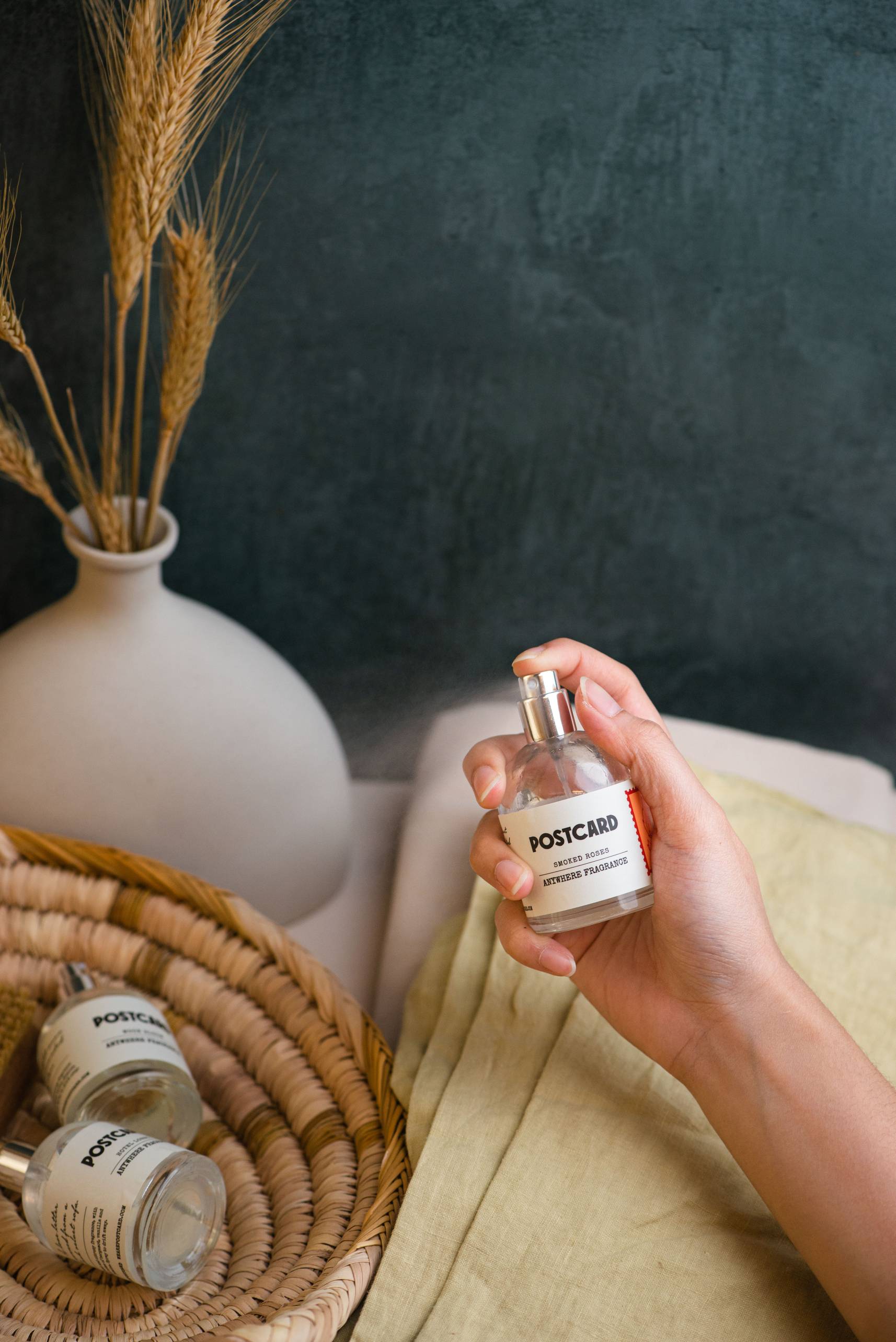 A hand holds a 50ml Postcard Anywhere Fragrance (Smoked Roses) bottle near two similar bottles, a woven basket, a white vase with dried wheat stems, and a light yellow cloth—all set against a dark background.