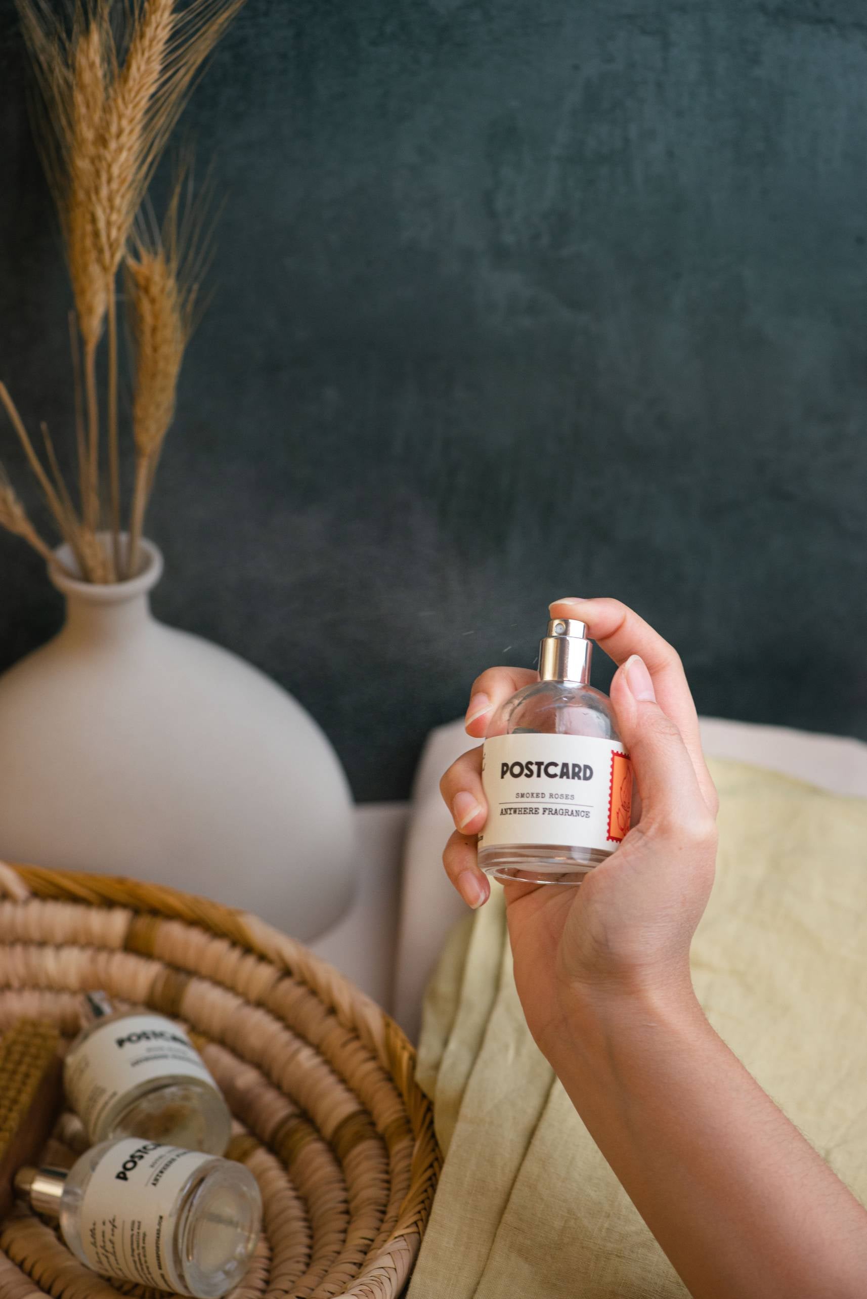 A hand holds a bottle of Postcard Anywhere Fragrance, 50ml - Smoked Roses, ready to spray. Two more identical Postcard bottles rest in a basket next to a white vase with dried wheat on a dark green background. Experience its unique smoked rose scent notes.