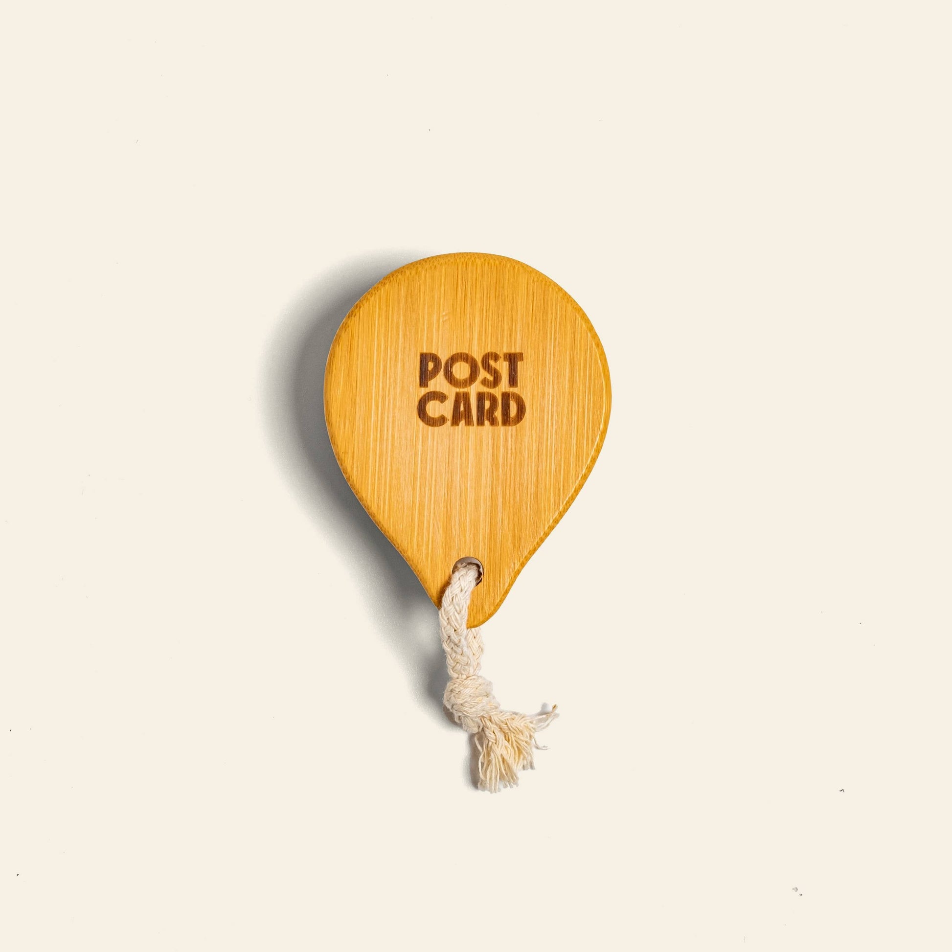 The Postcard Bamboo Scalp Massager Brush - Natural by Postcard is made from natural bamboo, with POST CARD engraved, and features a white knotted string, displayed against a plain light background.
