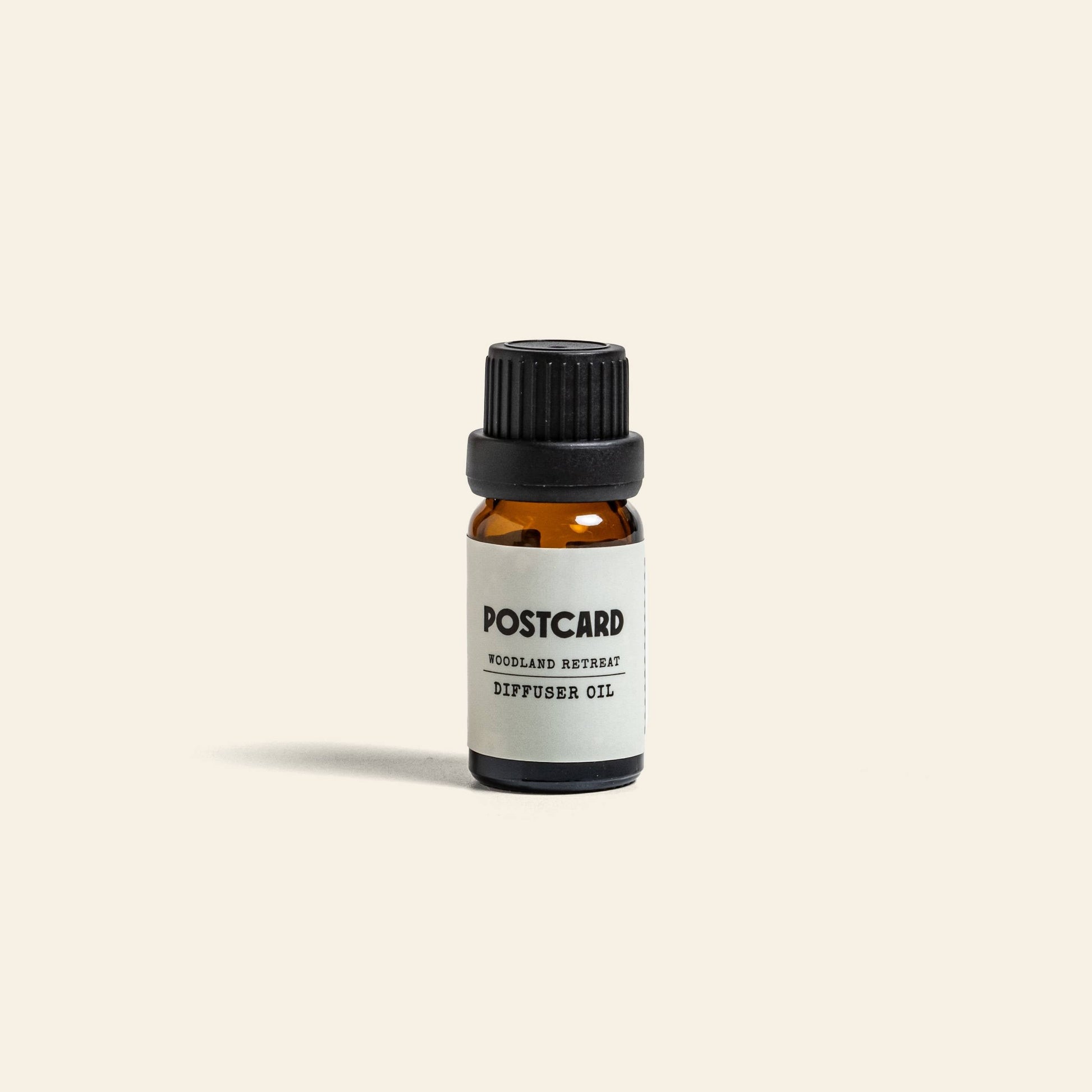 A small amber glass bottle with a black screw cap and white label reads “Postcard Diffuser Oil, 10g - Woodland Retreat” by Postcard, standing upright on a light beige background—infused with soothing forest-inspired scents.