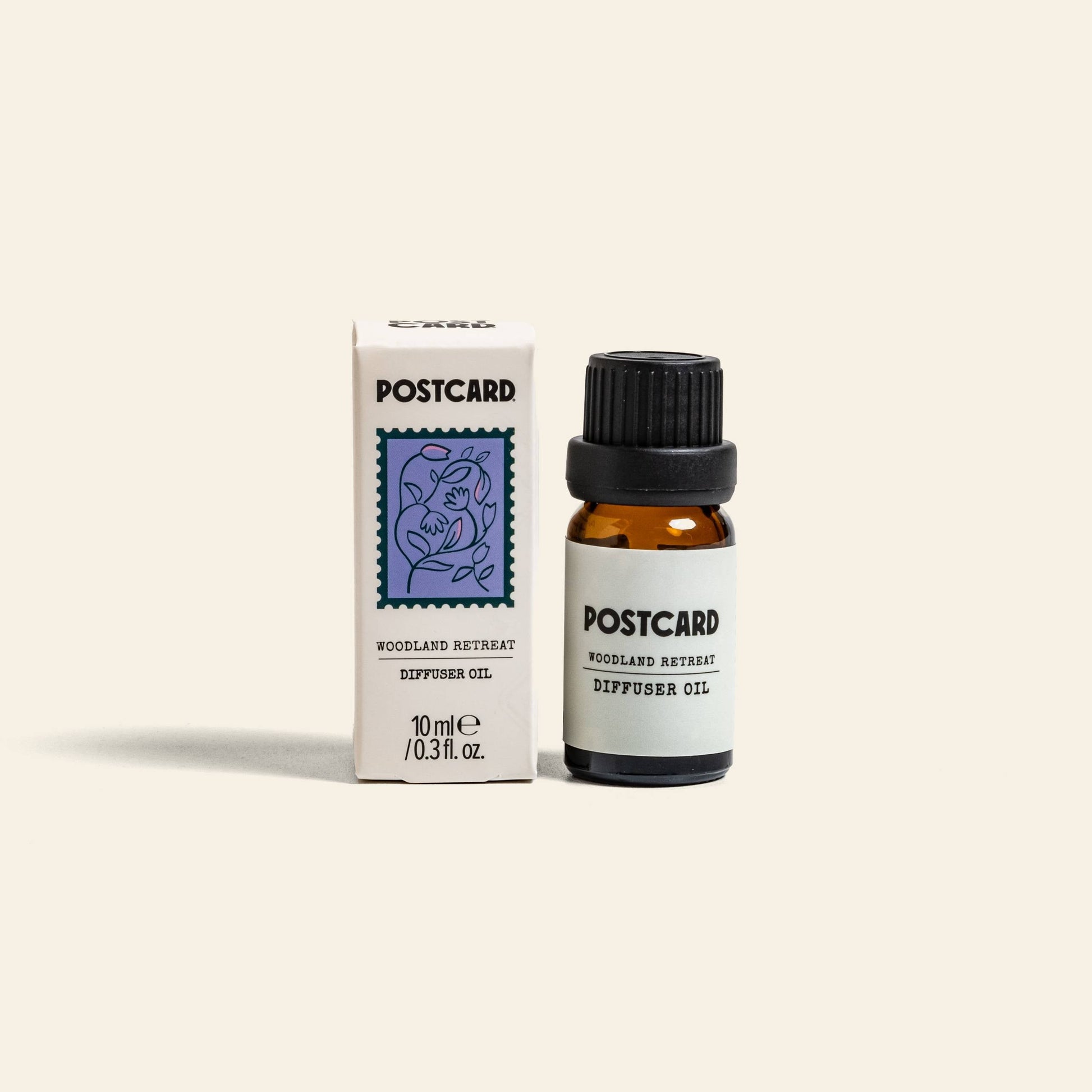 A 10g bottle of Postcard Diffuser Oil in the Woodland Retreat scent sits beside its box, which features a floral stamp illustration. The small amber bottle offers a calming forest elixir from the Postcard brand.