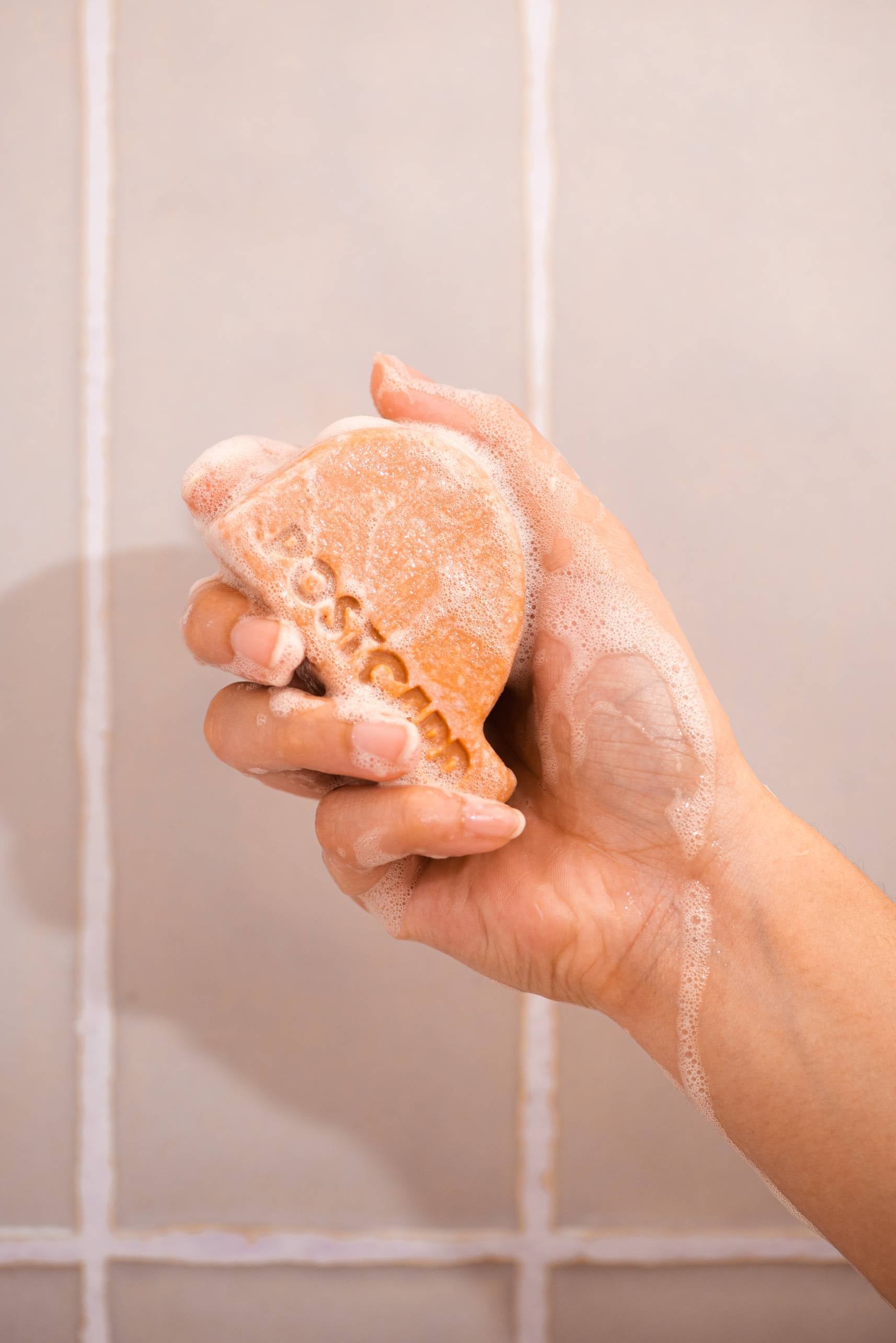 A hand holds the Postcard Head-To-Toe Bar, 100g - Citrus Circus, a sulfate-free, shell-shaped orange soap lathered with suds against tiled walls. This bar by Postcard gently pampers and energises your skin.