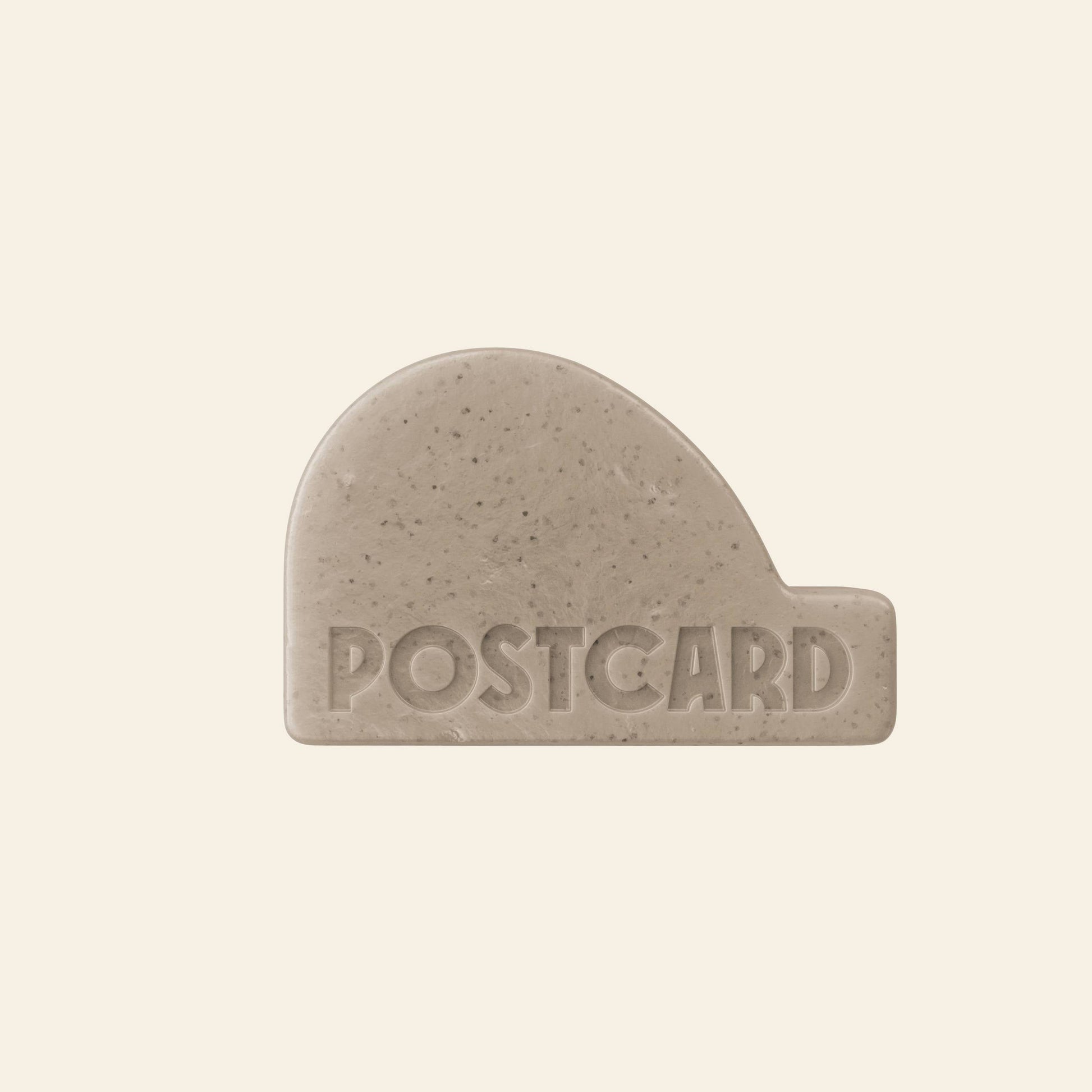 The Postcard Head-To-Toe Bar, 100g - Coffee Spot by Postcard is a beige, uniquely shaped exfoliating bar with POSTCARD engraved on it, set against a plain light background.