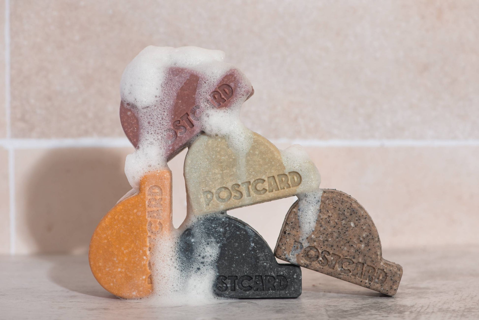 Five colorful Postcard Head-To-Toe Bars (100g, Coffee Spot) are stacked on tiles with white foam—ideal for your next coffee-infused, sulfate-free cleansing or shampoo routine.