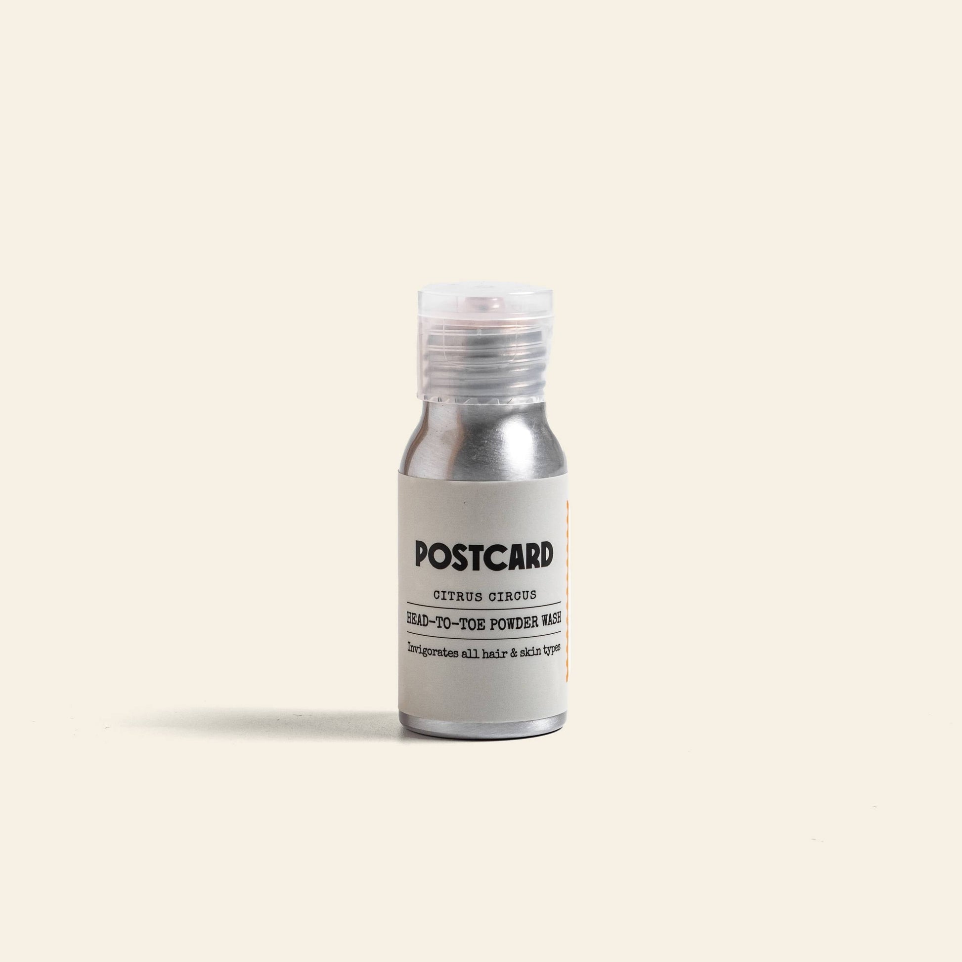 A 25g bottle of Postcard Head-To-Toe Powder Wash in Citrus Circus, a vitamin C-infused powder-to-foam cleanser, stands upright with a clear cap on a light beige background.
