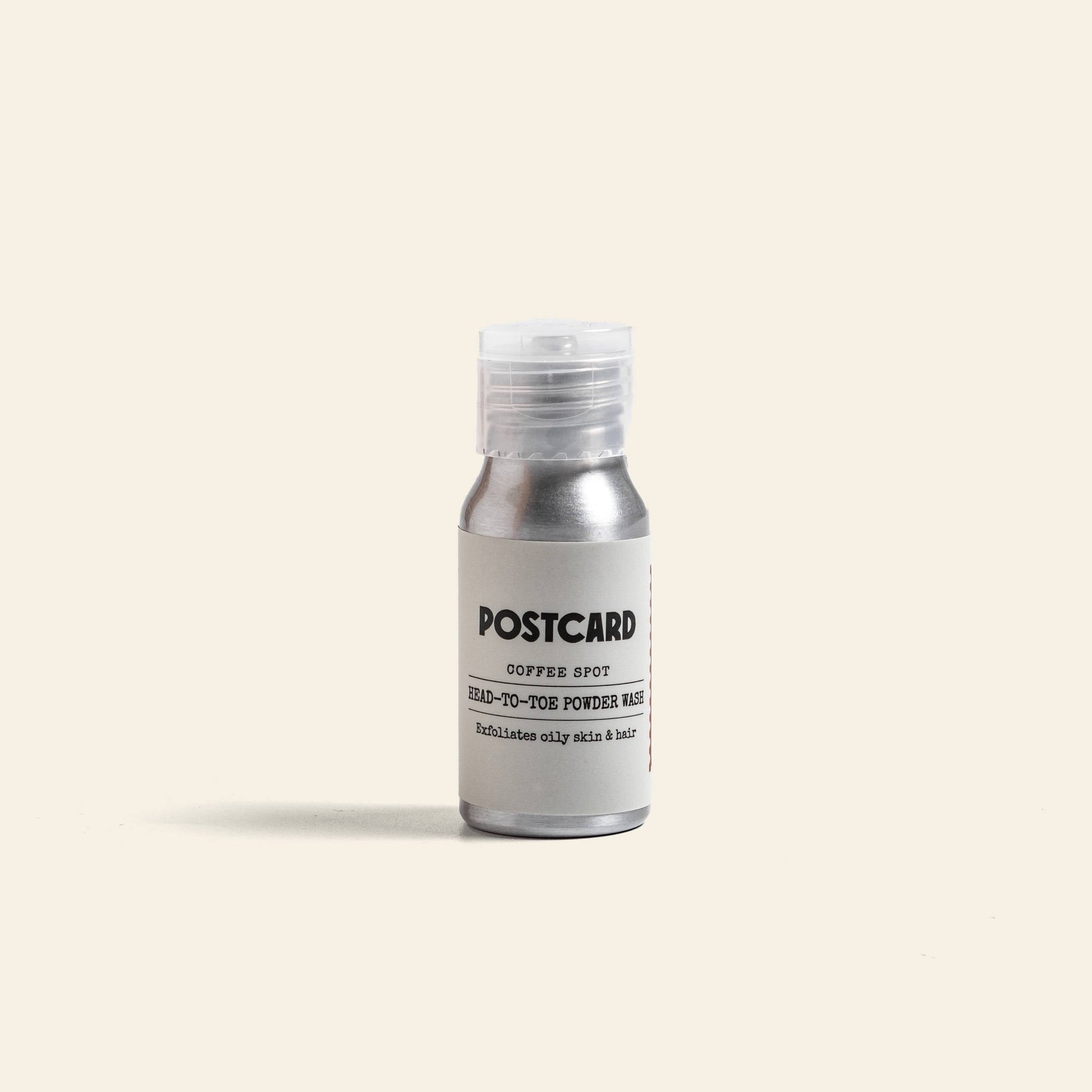 A small metallic bottle labeled “Postcard Head-To-Toe Powder Wash, 25g – Coffee Spot” stands upright on a light beige background, featuring a sulfate-free powder-to-foam cleanser formula from Postcard.