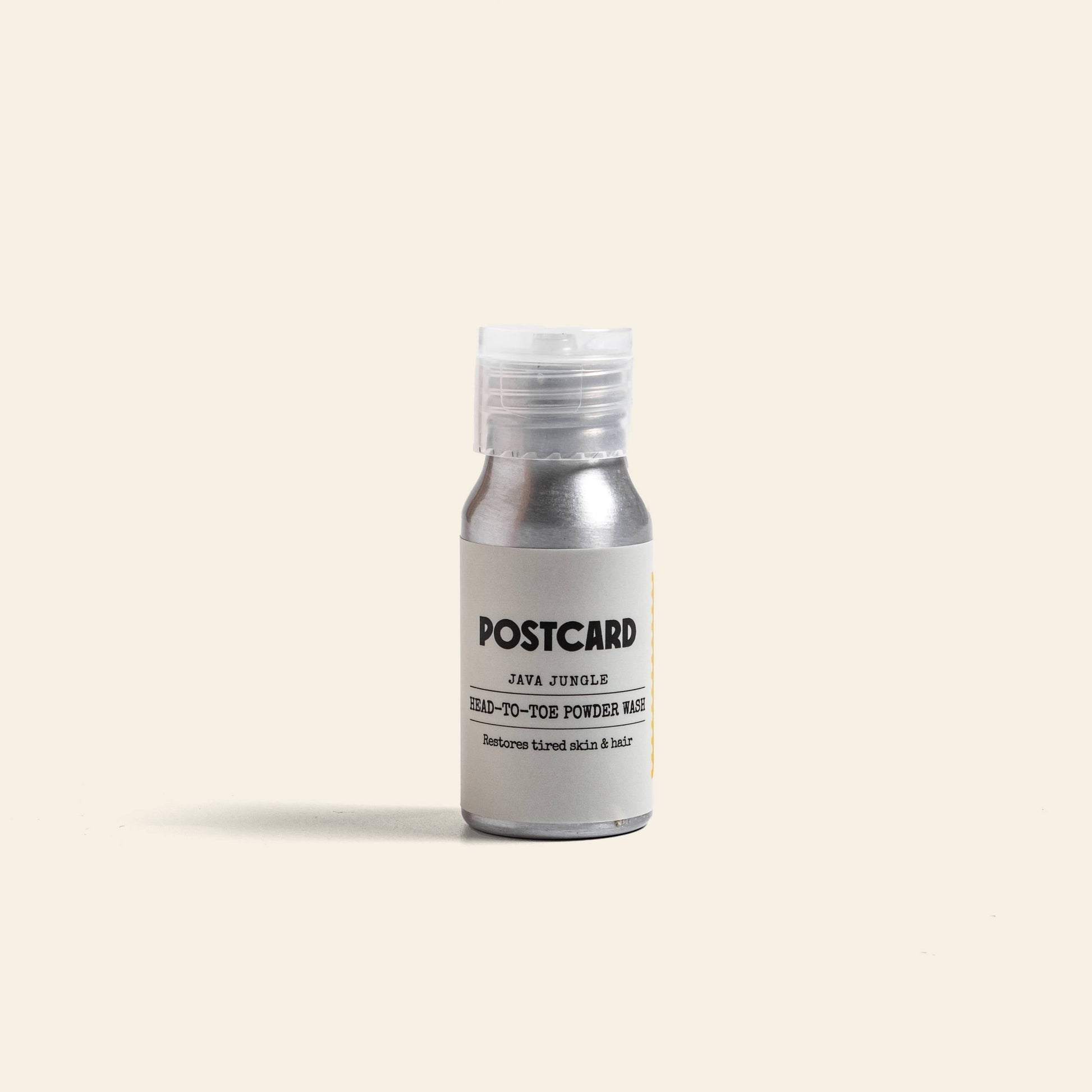 A small silver bottle of Postcard Head-To-Toe Powder Wash, 25g - Java Jungle, stands upright on a light beige background with a white cap; this sulfate-free powder wash features Stinging Nettle and is branded Postcard.