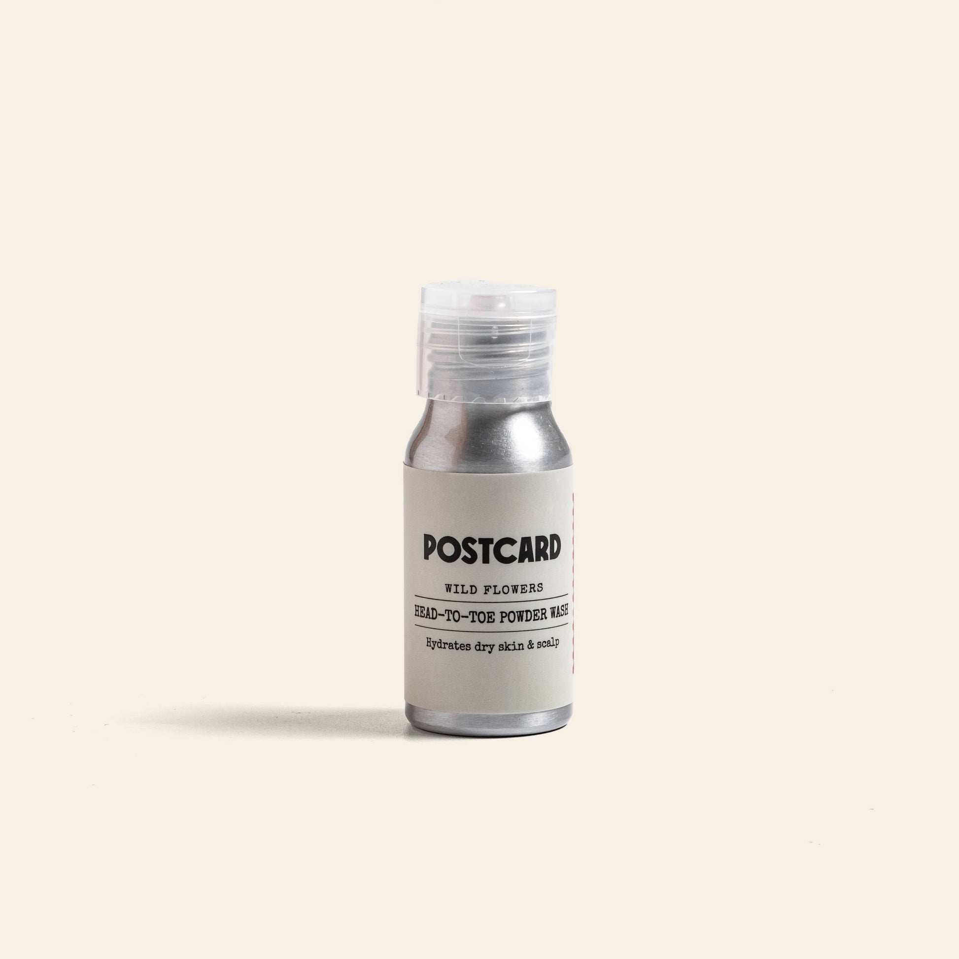 A small silver bottle with a clear cap labeled Postcard Head-To-Toe Powder Wash, 25g - Wild Flowers by Postcard, a sulfate-free cleanser, stands upright against a plain beige background.