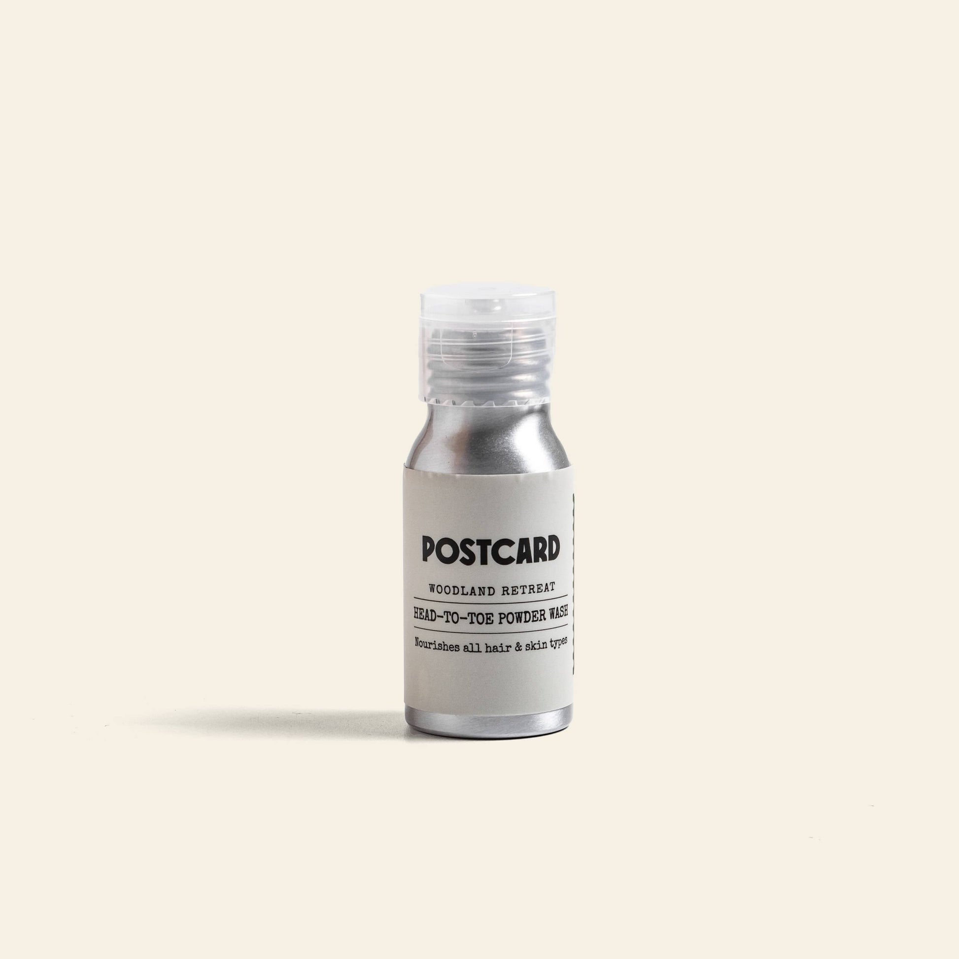 A small silver bottle with a clear cap, labeled Postcard Head-To-Toe Powder Wash, 25g - Woodland Retreat by Postcard, stands upright on a plain background, ready to reveal its herbal powder-to-foam cleanser.