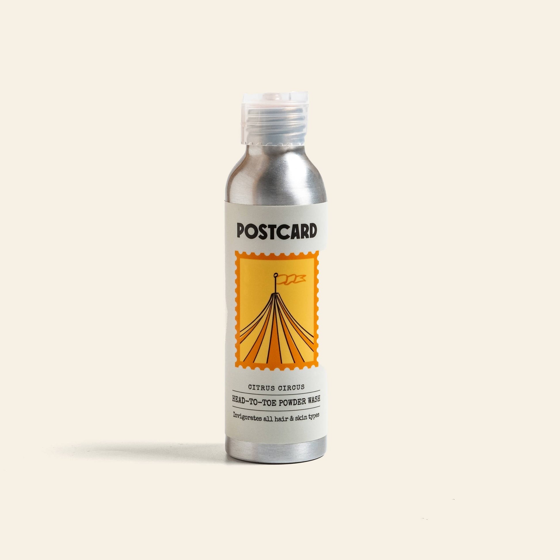 A silver bottle labeled Postcard Head-To-Toe Powder Wash, 80g - Citrus Circus, features a yellow circus tent in a stamp-like border and holds a citrus-infused powder-to-foam cleanser with vitamin C for refreshing hair and skin care.