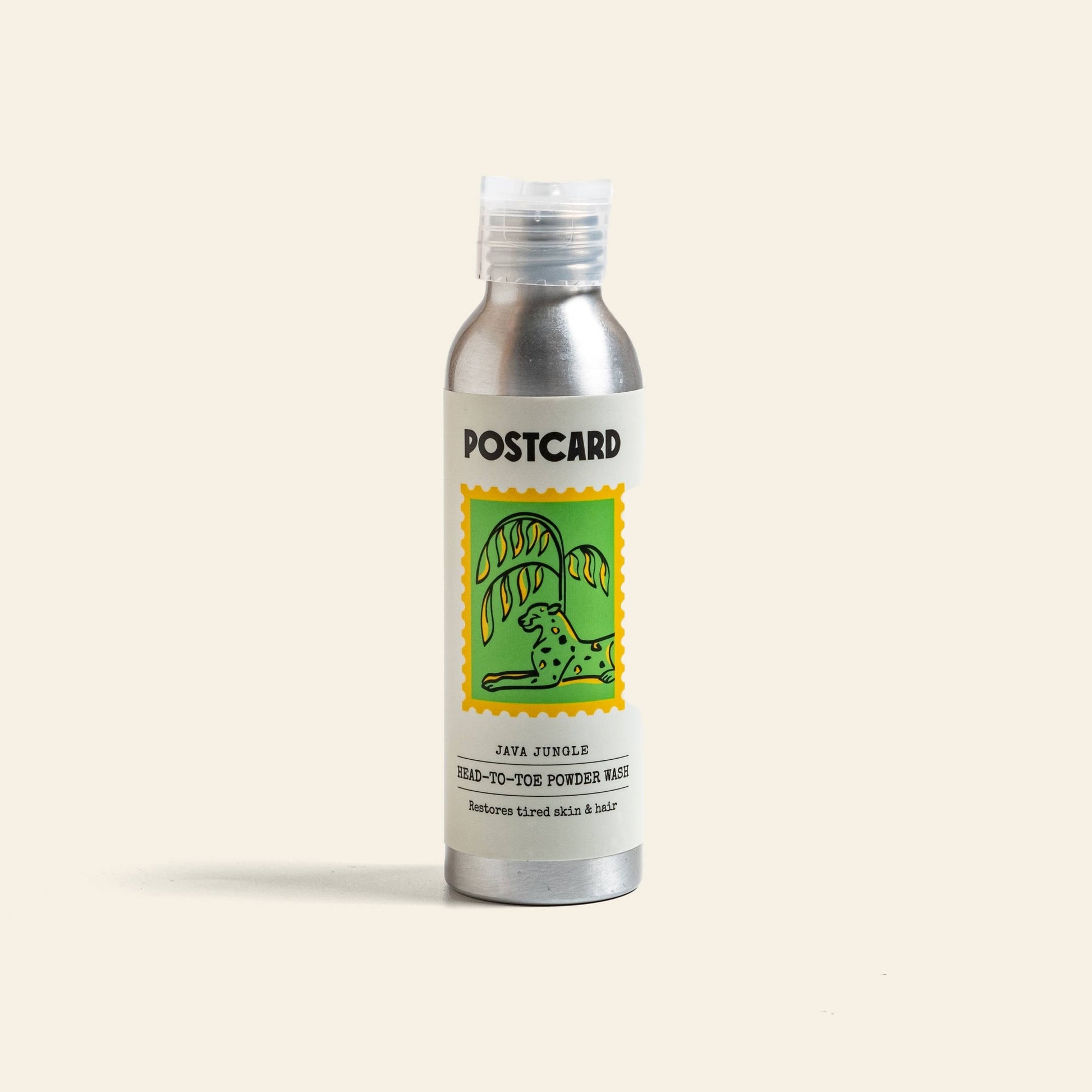 A silver bottle labeled Postcard features a colorful spotted animal beneath palm leaves. Below: Postcard Head-To-Toe Powder Wash, 80g – Java Jungle, a sulfate-free powder wash that revitalizes tired skin & hair and aids hair loss recovery.