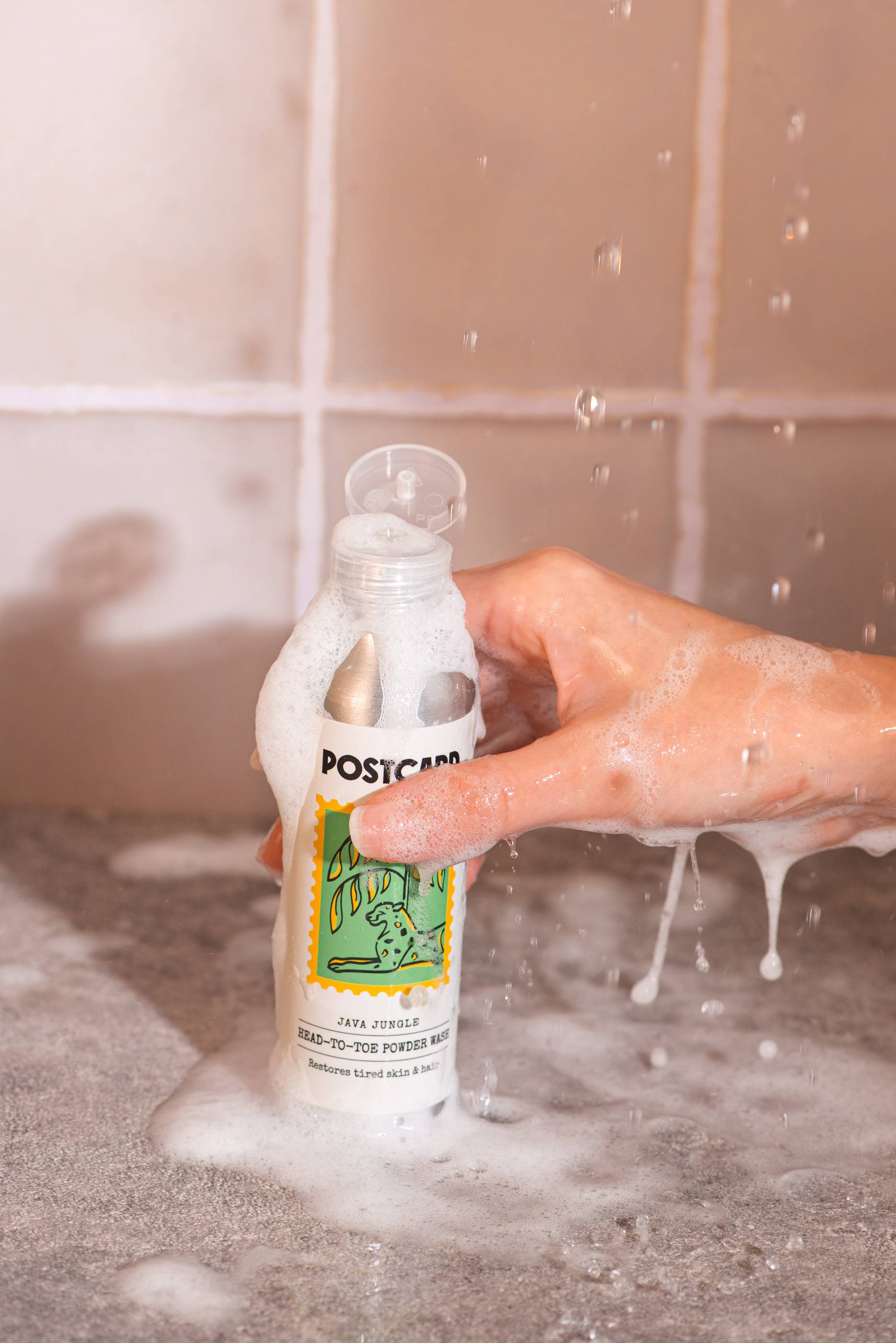 A hand holds a foamy bottle of Postcard Head-To-Toe Powder Wash, 80g - Java Jungle by Postcard under running water, with soap suds and droplets on a tiled surface.