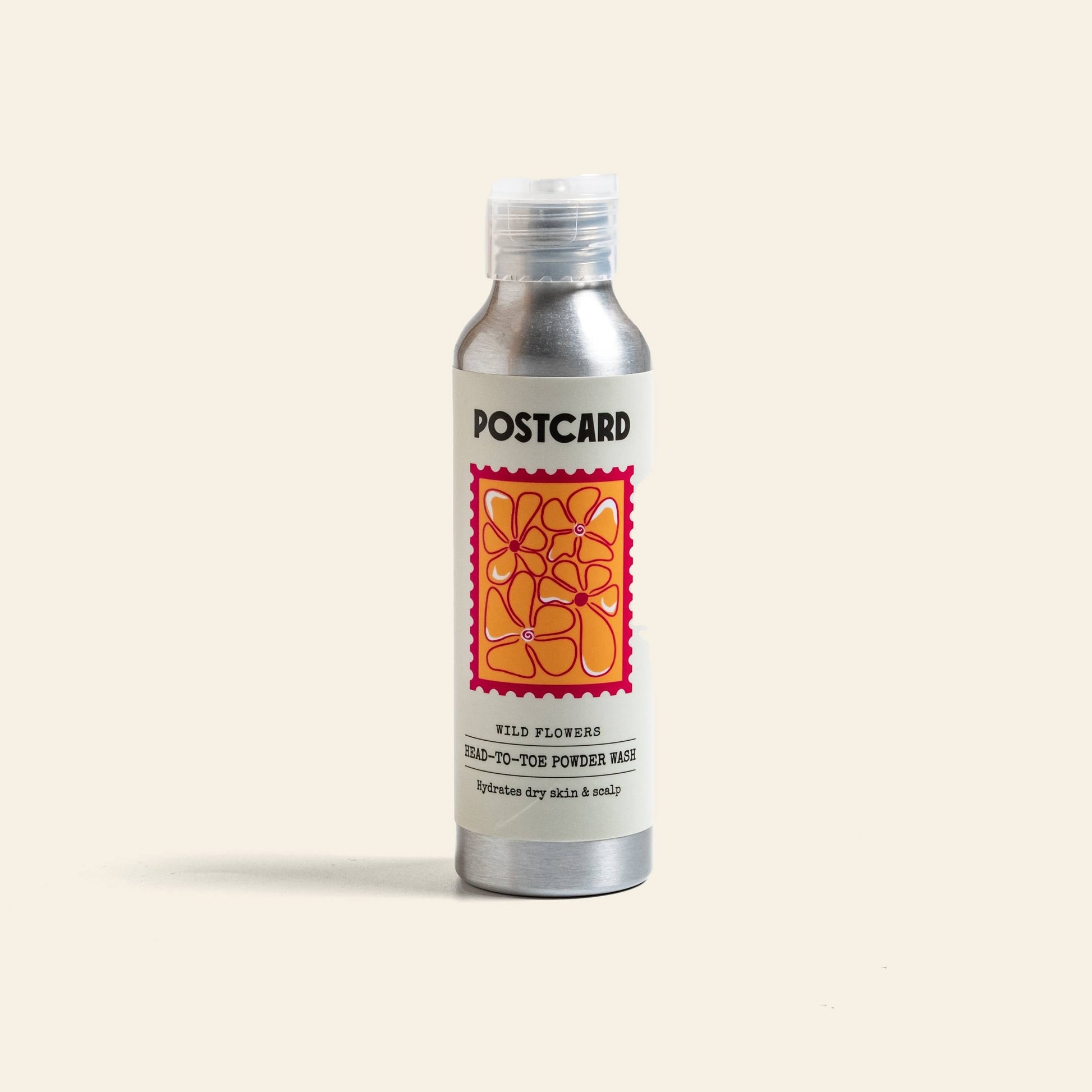 A silver 80g bottle by Postcard labeled “Postcard Head-To-Toe Powder Wash - Wild Flowers” features an abstract orange-yellow flower and a gentle, sulfate-free cleanser for hydrating dry skin and scalp.
