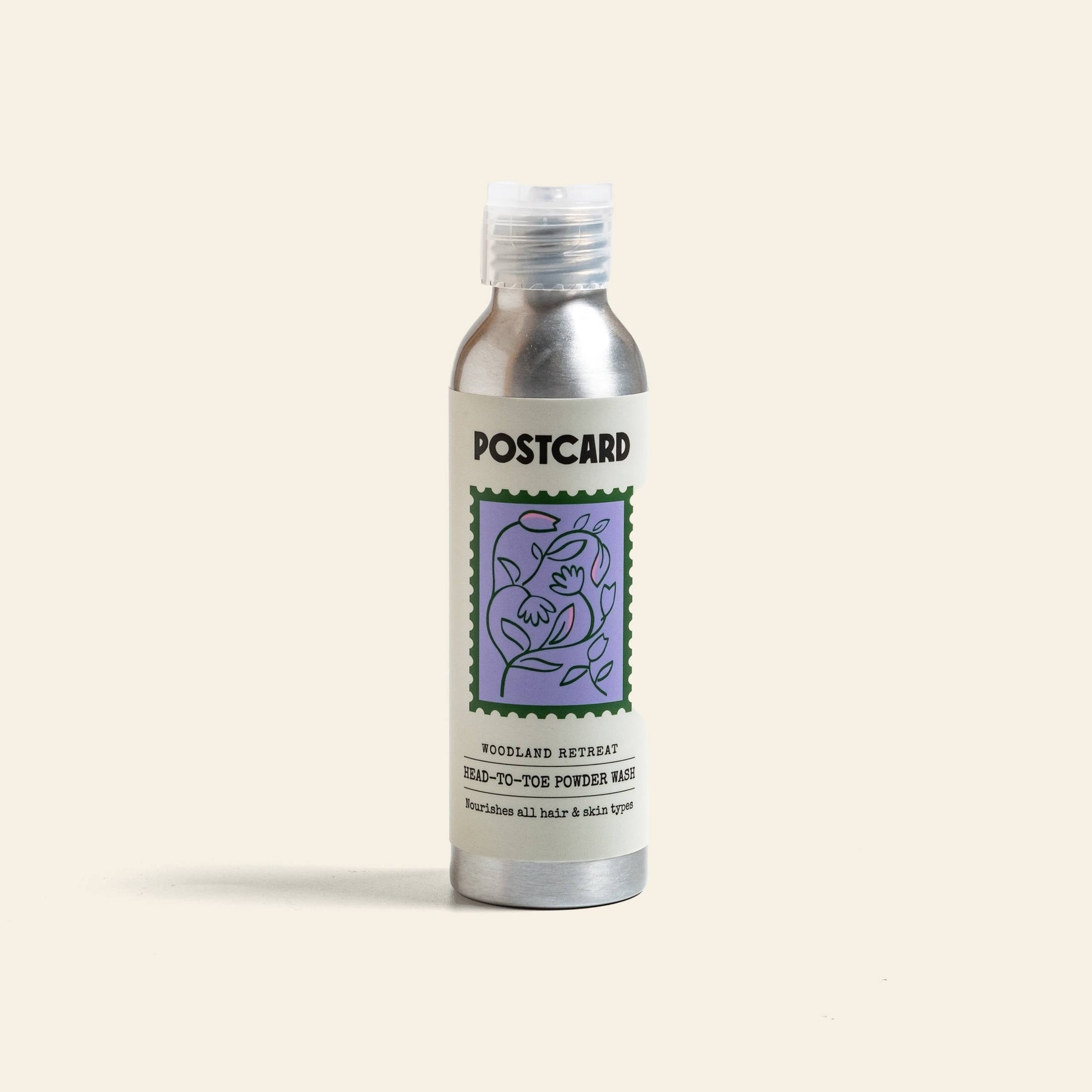 A silver 80g Postcard Head-To-Toe Powder Wash bottle, Woodland Retreat scent, with a purple floral design, stands upright on a plain background, evoking a luxurious spa experience.