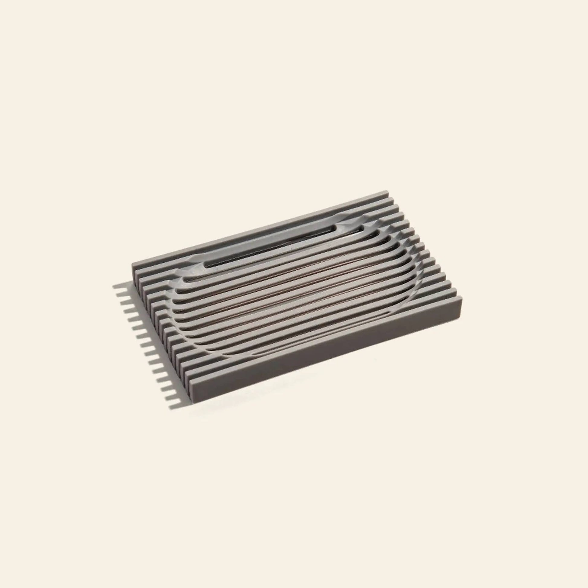 The Postcard Lattice Soap Dish by Postcard is a rectangular, dark gray soap dish made from recyclable resin with parallel slats and a central oval, perfect for a zero-waste lifestyle on any plain light surface.
