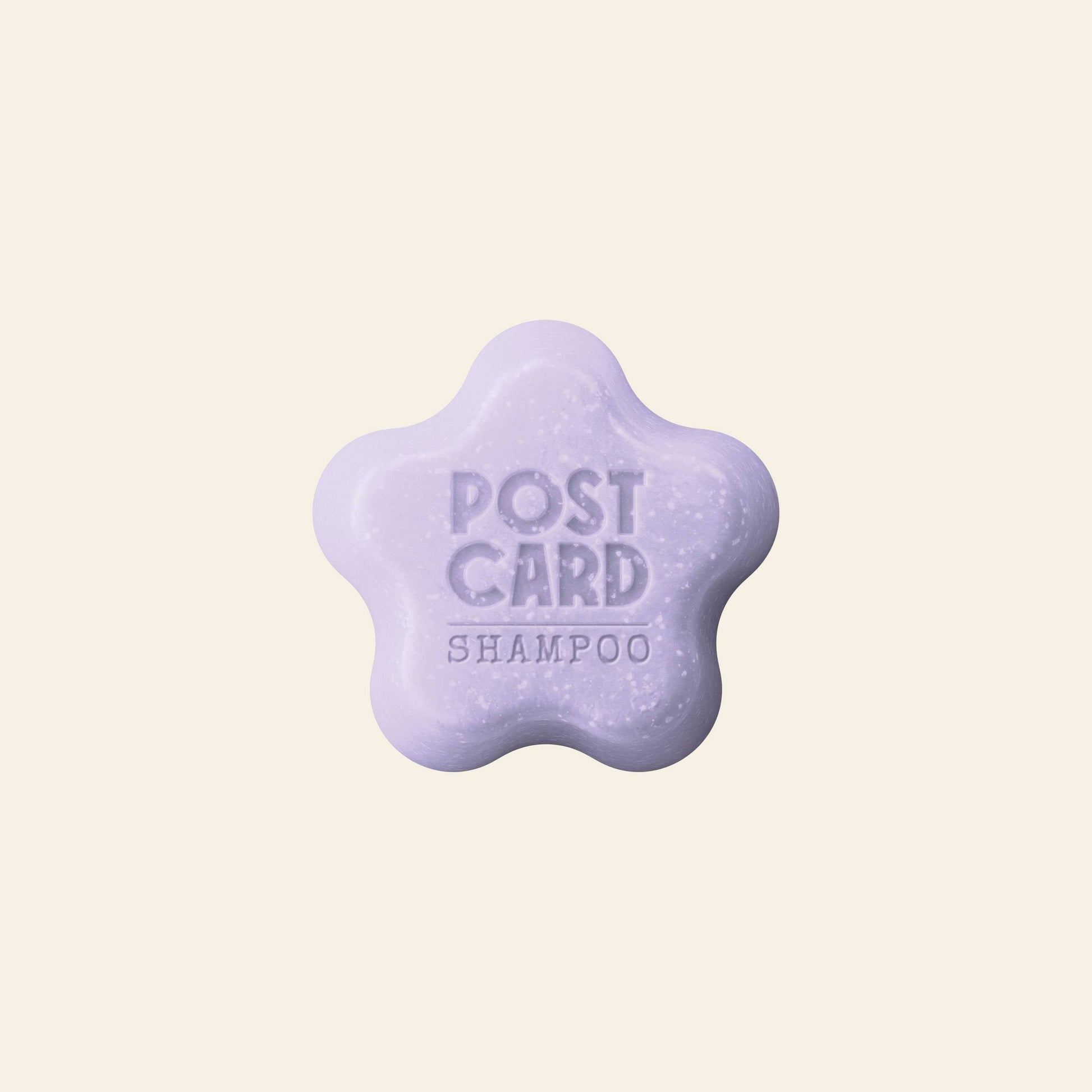 A light purple, flower-shaped Postcard PET Shampoo Bar (60g) by Grooming Room with POST CARD SHAMPOO embossed, made from natural ingredients, displayed on a plain cream background.