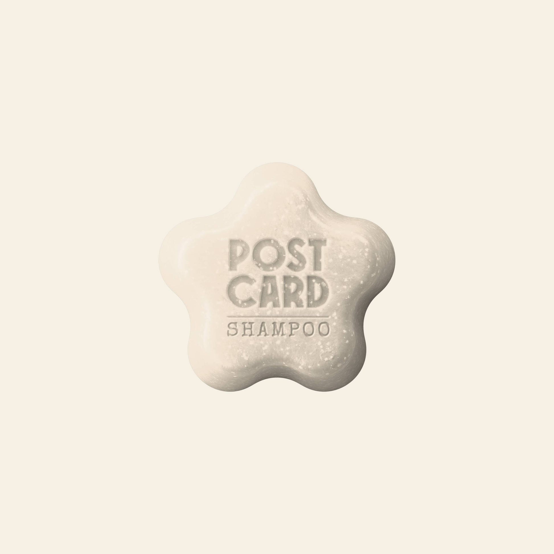 A flower-shaped Postcard Shampoo Bar, 60g – Clear Skies, embossed with POSTCARD SHAMPOO, rests on a light beige background. This sulfate-free bar from Postcard is ideal for sensitive scalps needing gentle, effective cleansing.