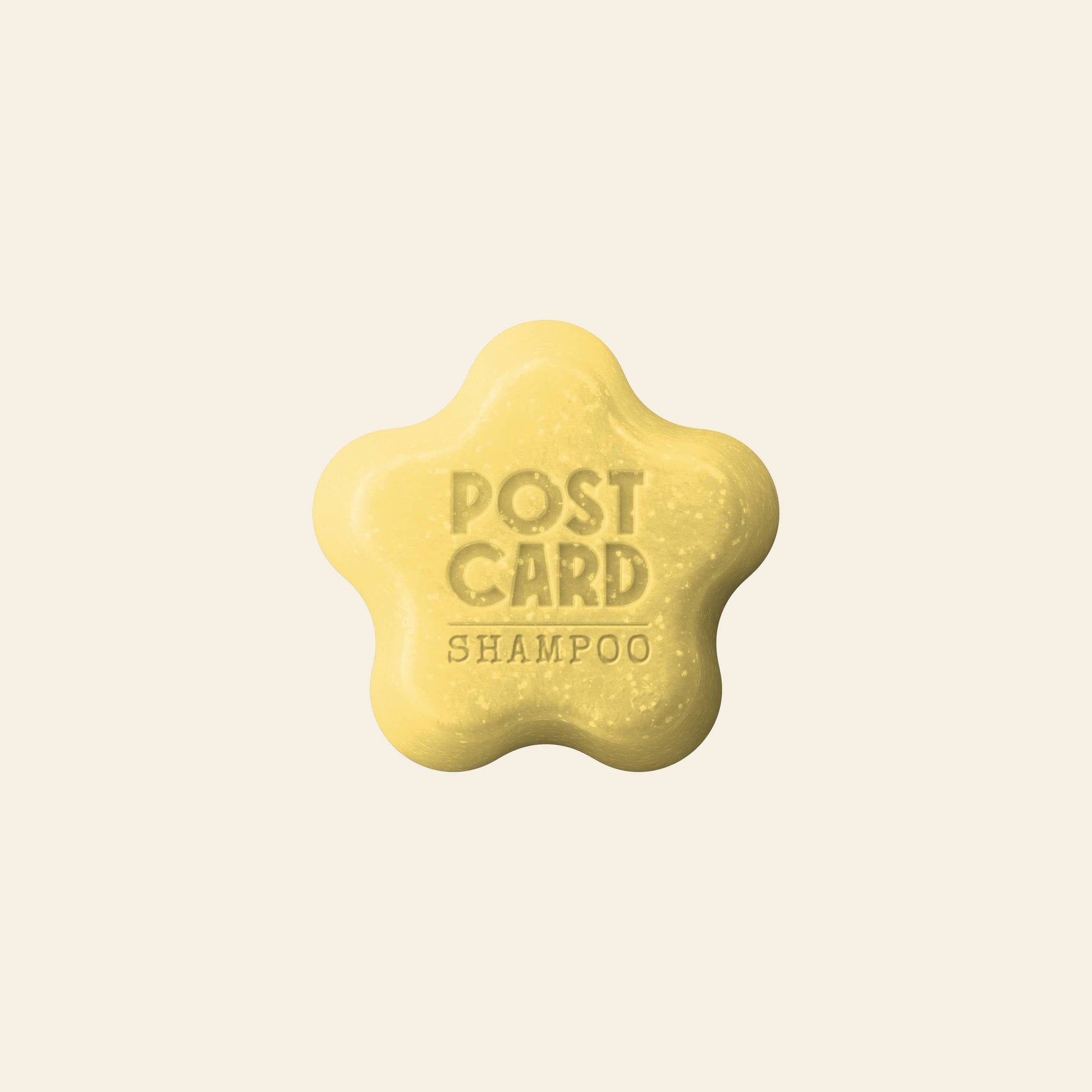 The Postcard Coconut Beach Shampoo Bar (60g) is a yellow, flower-shaped sulfate-free bar, made with raw virgin coconut oil and embossed with POST CARD SHAMPOO. It leaves hair nourished and refreshed. Set against a plain beige background.