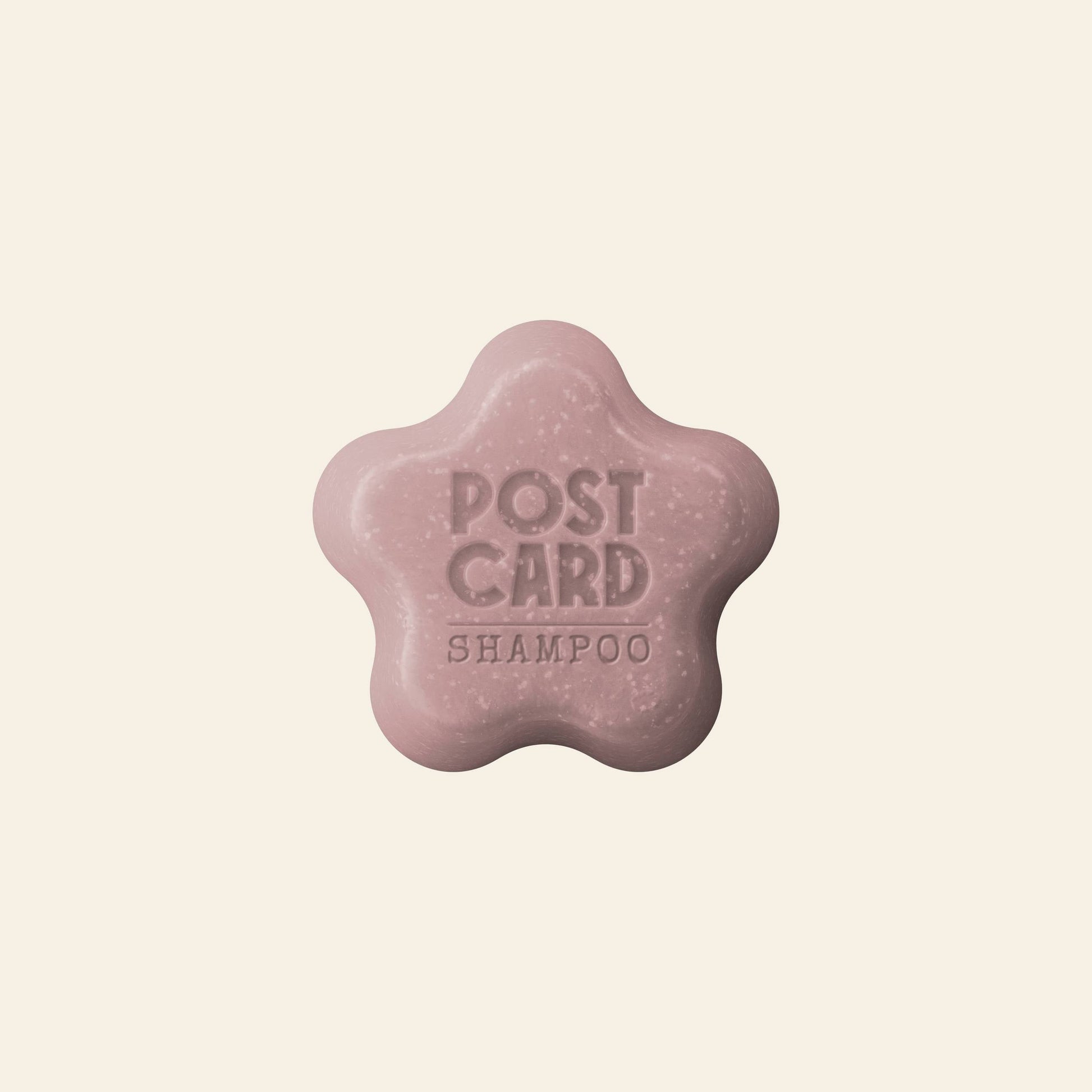 The Postcard Shampoo Bar, 60g – Wild Flowers, by Postcard, is a pink, flower-shaped bar embossed with POST CARD SHAMPOO, featuring a sulfate-free formula for hydration, shown on a plain light background.
