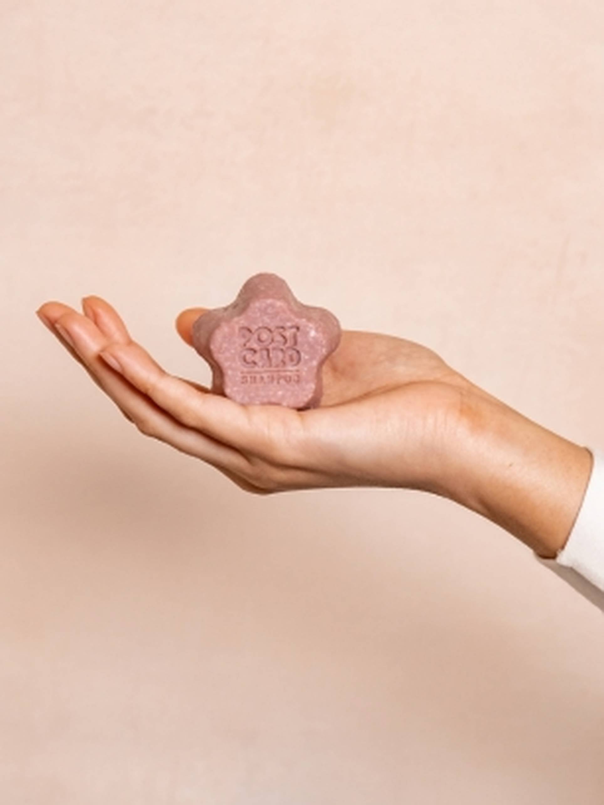 A hand holds the Postcard Shampoo Bar, 60g - Wild Flowers by Postcard, a pink star-shaped bar embossed with POST CARD SHAMPOO, offering hydration against a soft beige background.