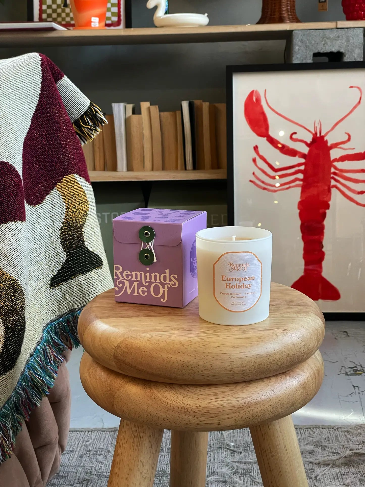 A round wooden stool holds a purple Reminds Me Of box and a Reminds Me Of European Holiday Candle, 10oz. Nearby, a patterned blanket hangs with books and red lobster artwork in the background.