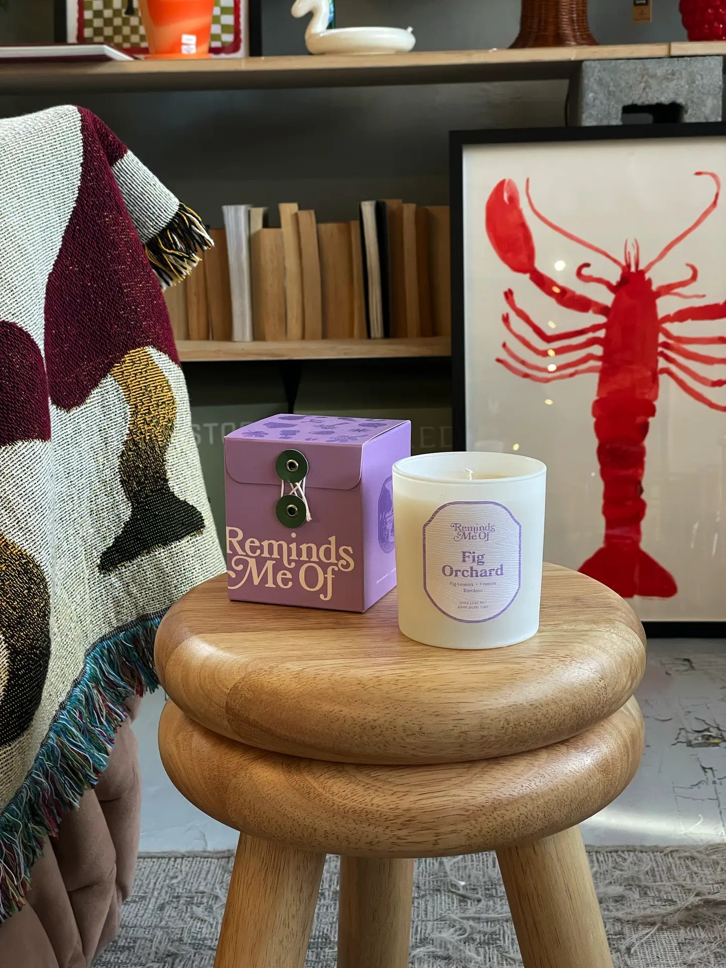 A wooden stool displays a purple Reminds Me Of Fig Orchard Candle, 10oz by Reminds Me Of. A colorful blanket hangs nearby, with shelves and red lobster artwork visible in the background.