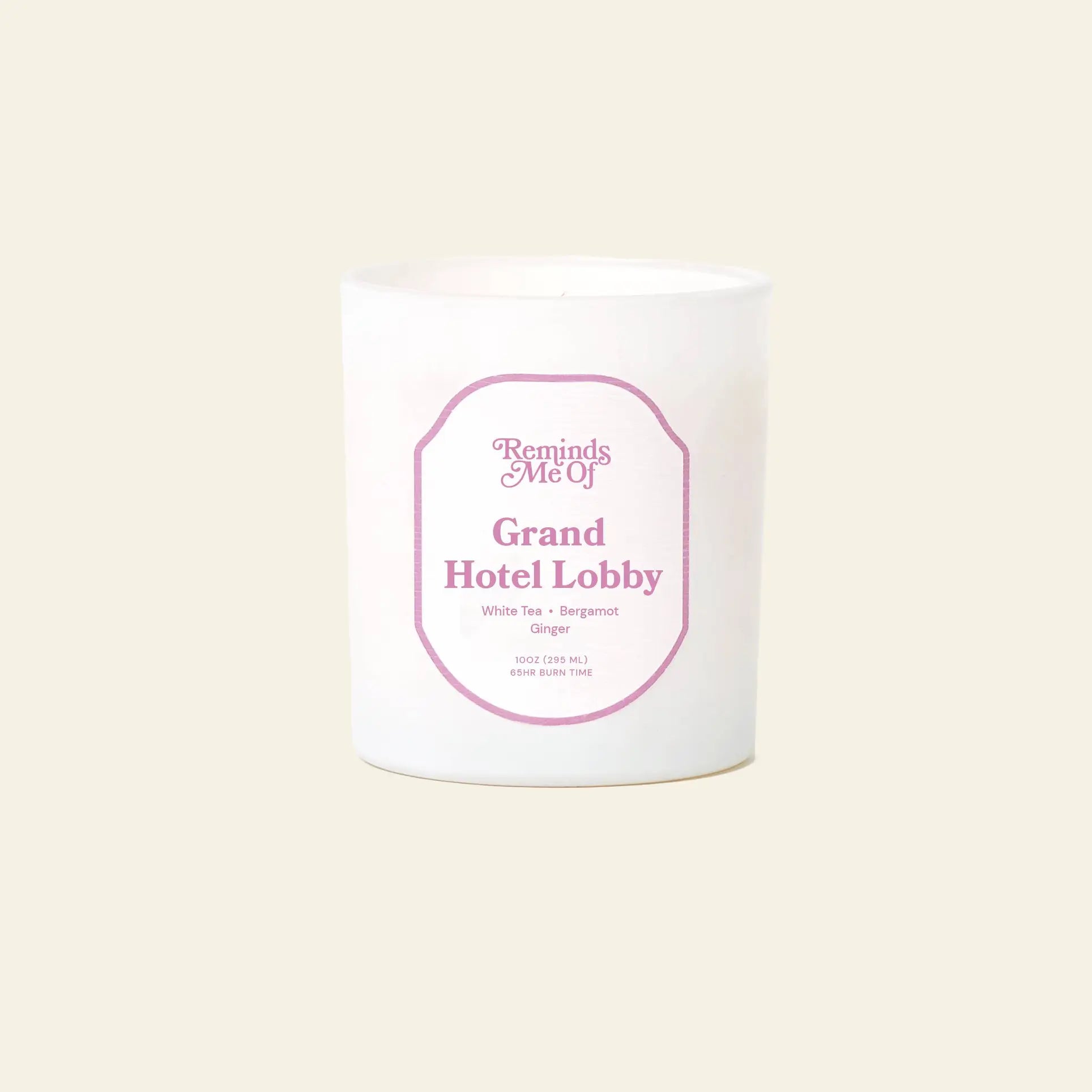 The Reminds Me Of Grand Hotel Lobby Candle, 10oz features uplifting scents of White Tea, Bergamot, and Ginger. This white candle with a pink label is set against a plain light beige background. Brand: Reminds Me Of.