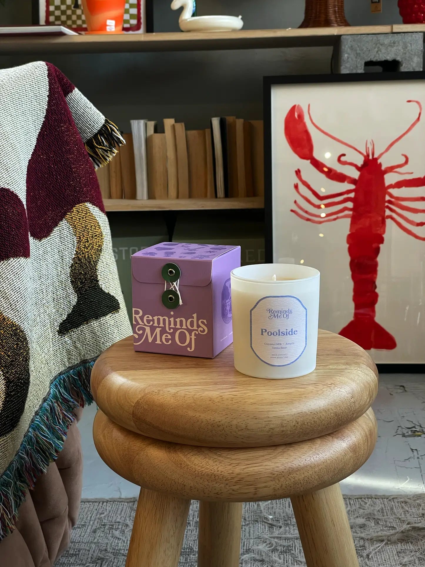 A wooden stool holds a Reminds Me Of Poolside Candle, 10oz, in purple and coconut milk scent, next to a patterned blanket and framed red lobster artwork on a bookshelf.