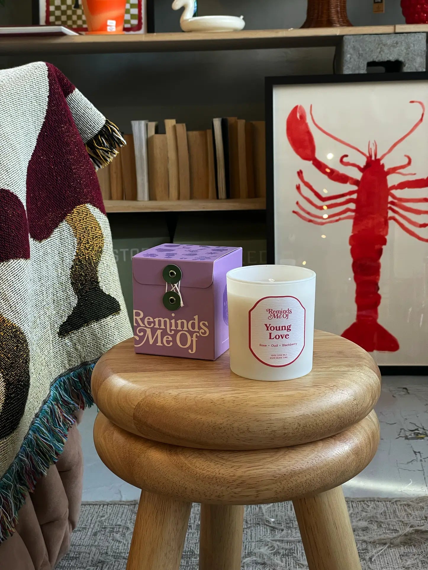 A 10oz Reminds Me Of Young Love Candle rests on a wooden stool in its purple box, surrounded by red lobster art, books, and a patterned blanket draped over furniture.