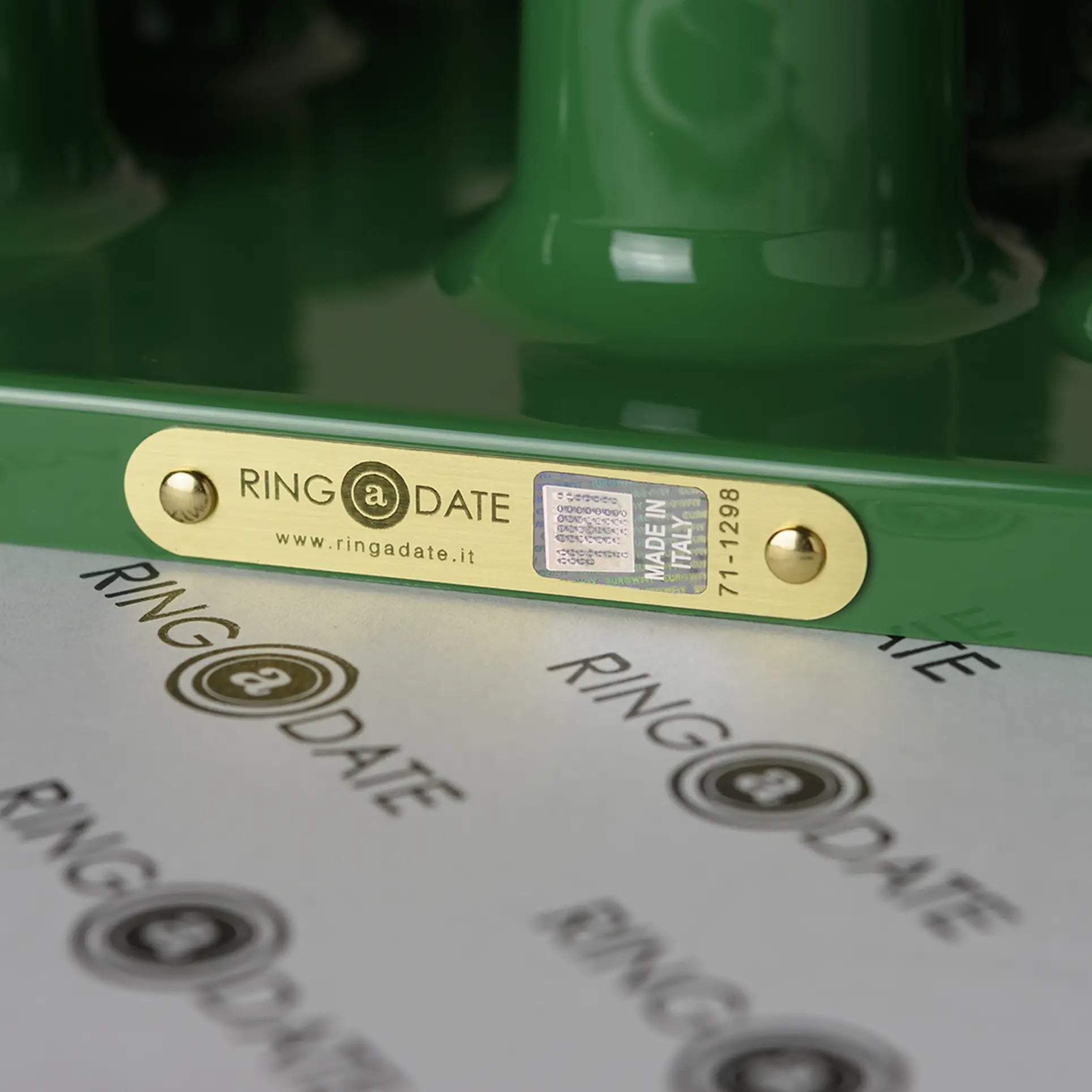 Close-up of a gold metal label on a green Ring A Date Wall Calendar by Ring-A-Date, showing RING @ DATE, a QR code, serial number, and MADE IN ITALY. The ABS plastic surface features repeating black and gold Ring-A-Date calendar logos.