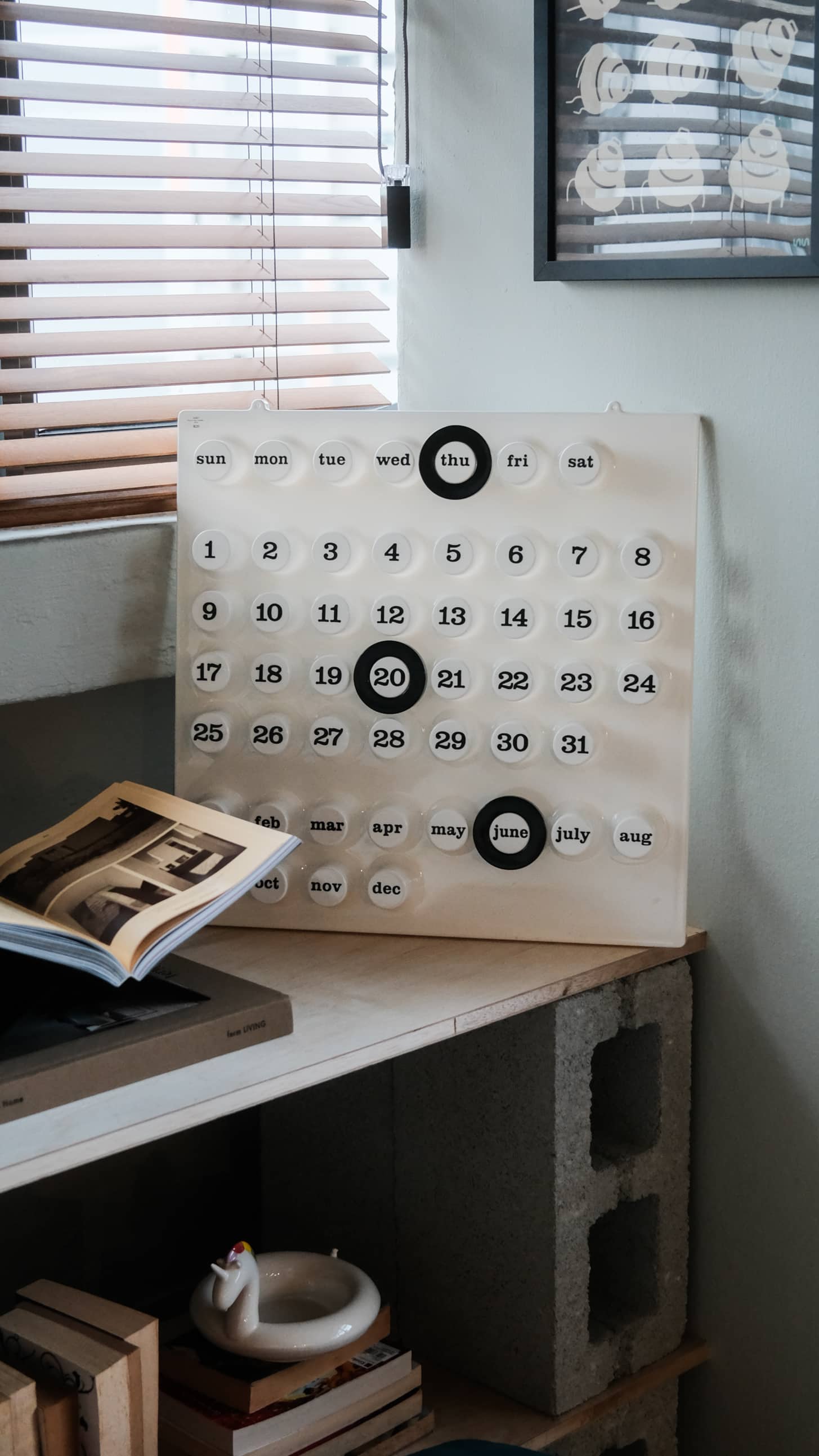 A modern, minimalist Ring A Date Wall Calendar by Ring-A-Date, made in Italy with movable circular markers, sits on a shelf near decor and an open book as sunlight streams through window blinds above.