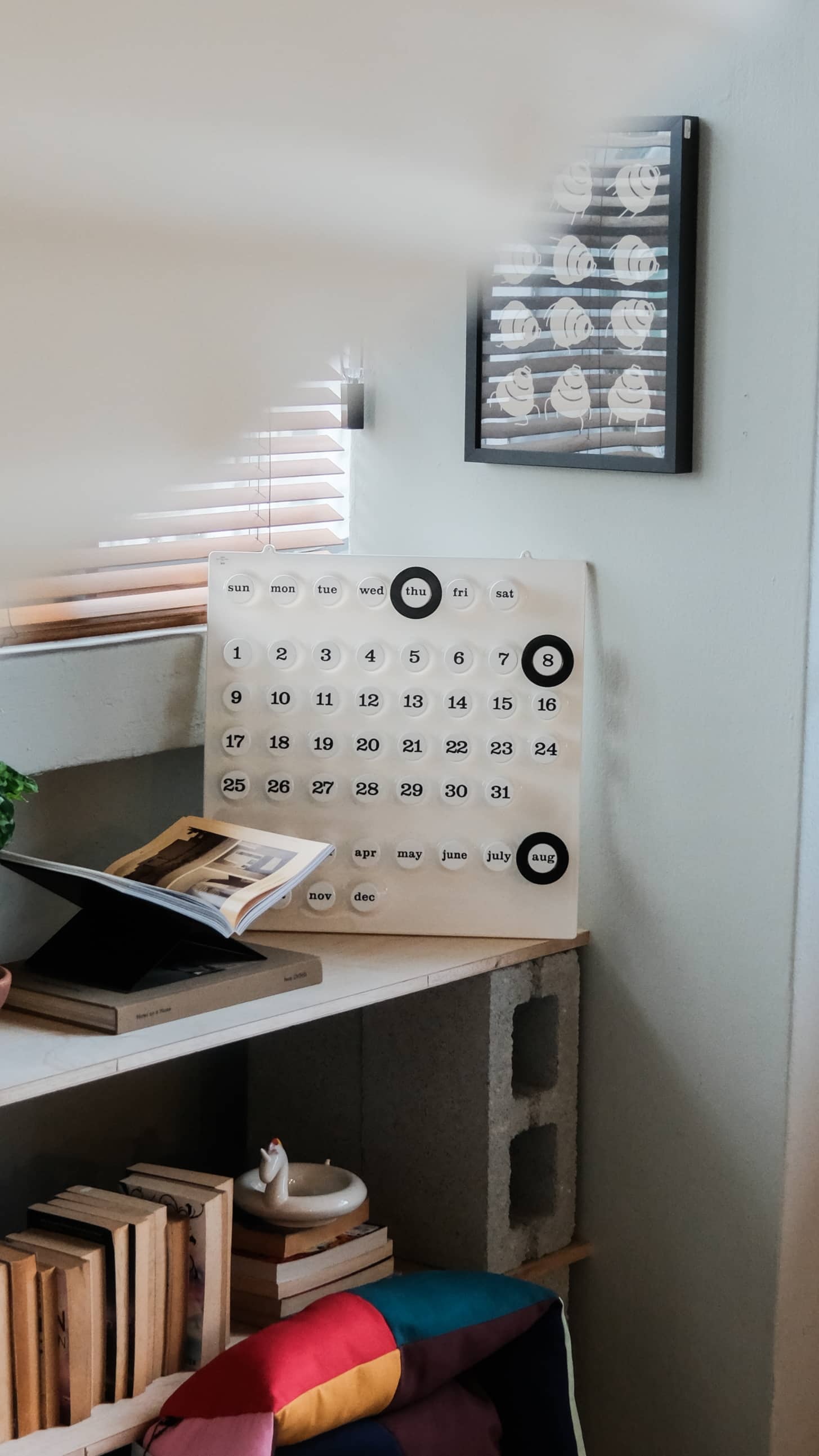 A minimalist room corner features the Ring-A-Date Wall Calendar by Ring-A-Date on a shelf, books stacked below, framed artwork above, and gentle light streaming through window blinds.