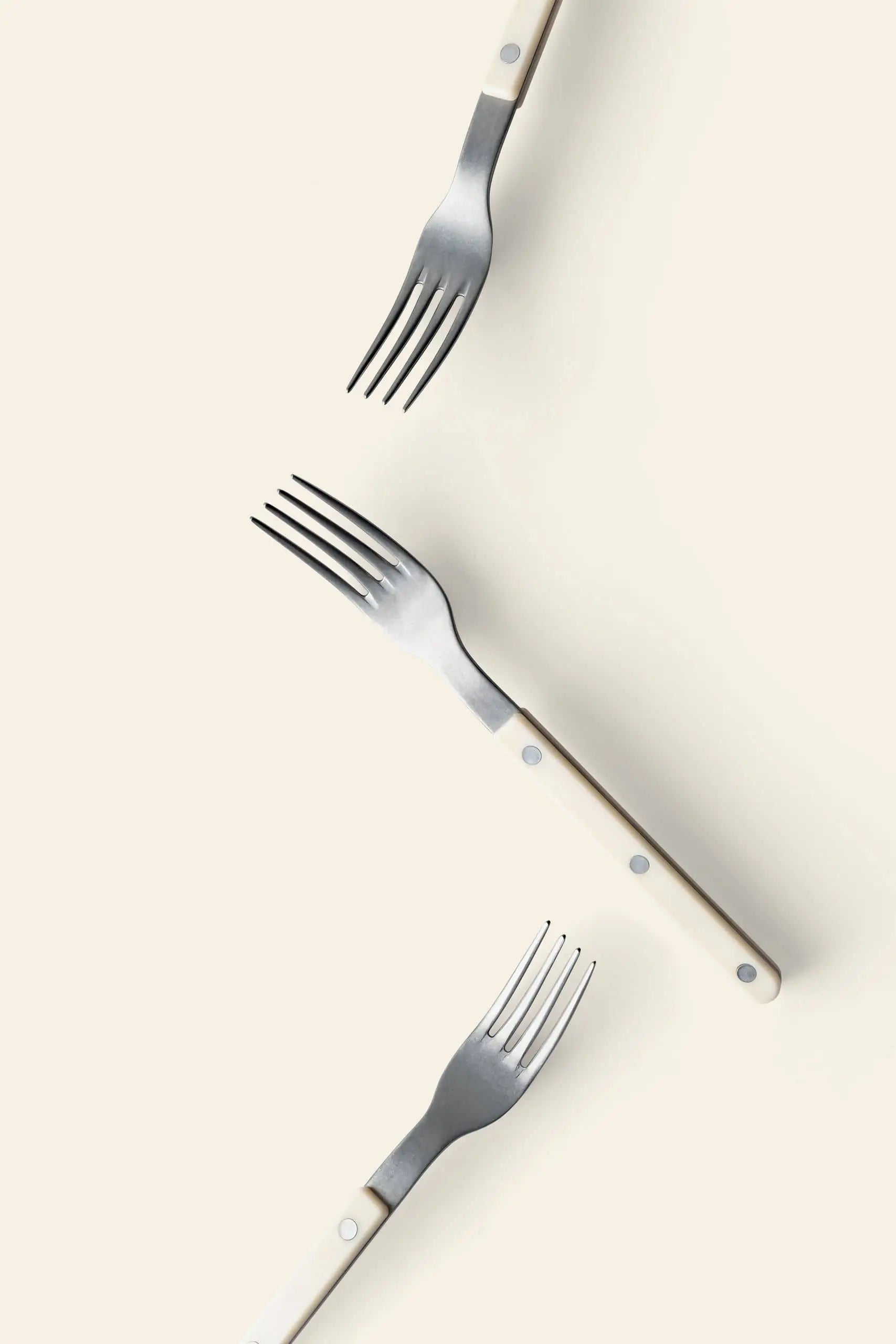 Three Sabre Bistrot Vintage Dinner Forks by Sabre, featuring white handles and stainless steel prongs, are arranged diagonally on a light cream background, creating a spaced display with prongs pointing in various directions.